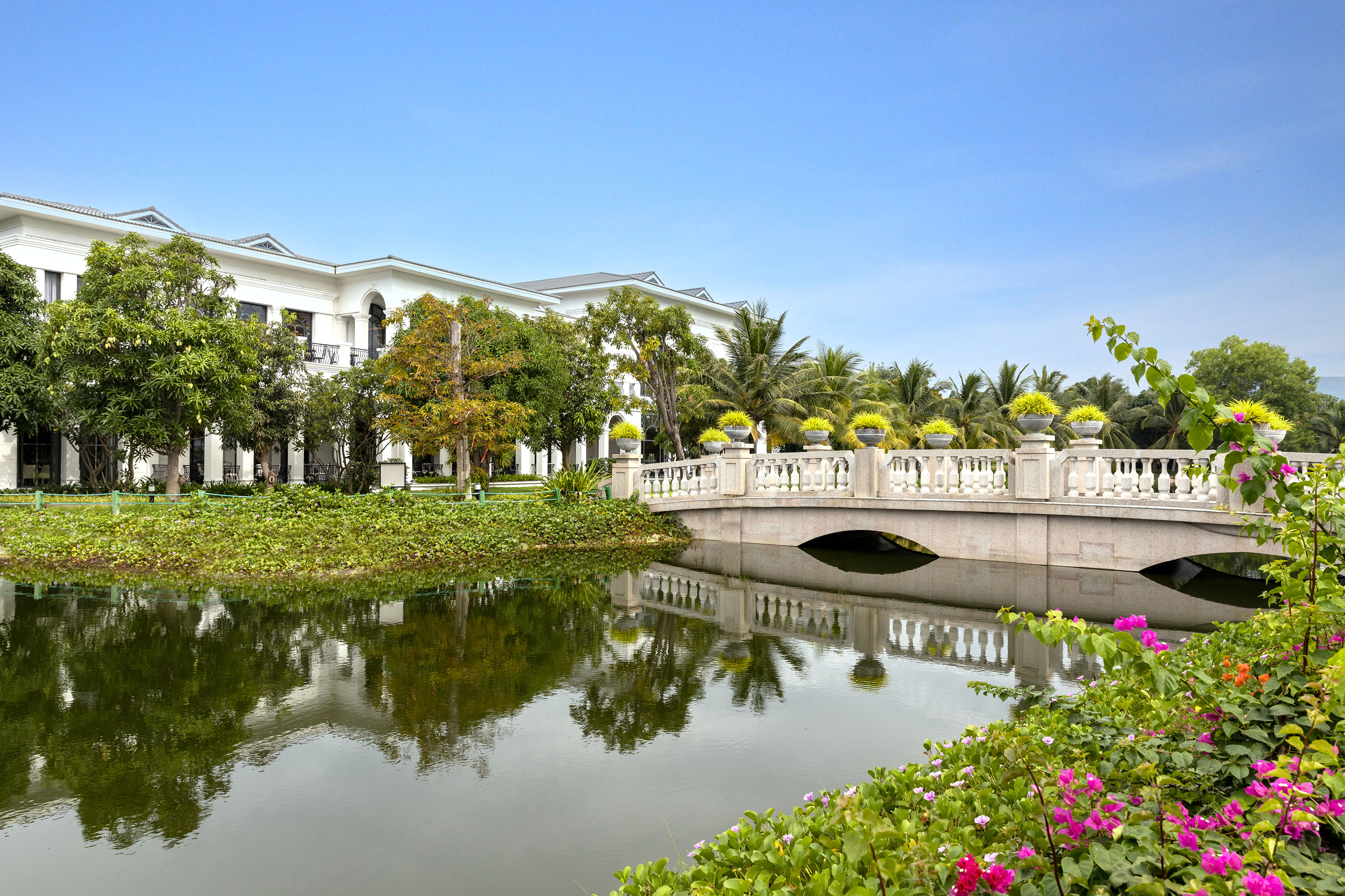 White resort building, stone bridge over canal, tropical gardens, blue sky.