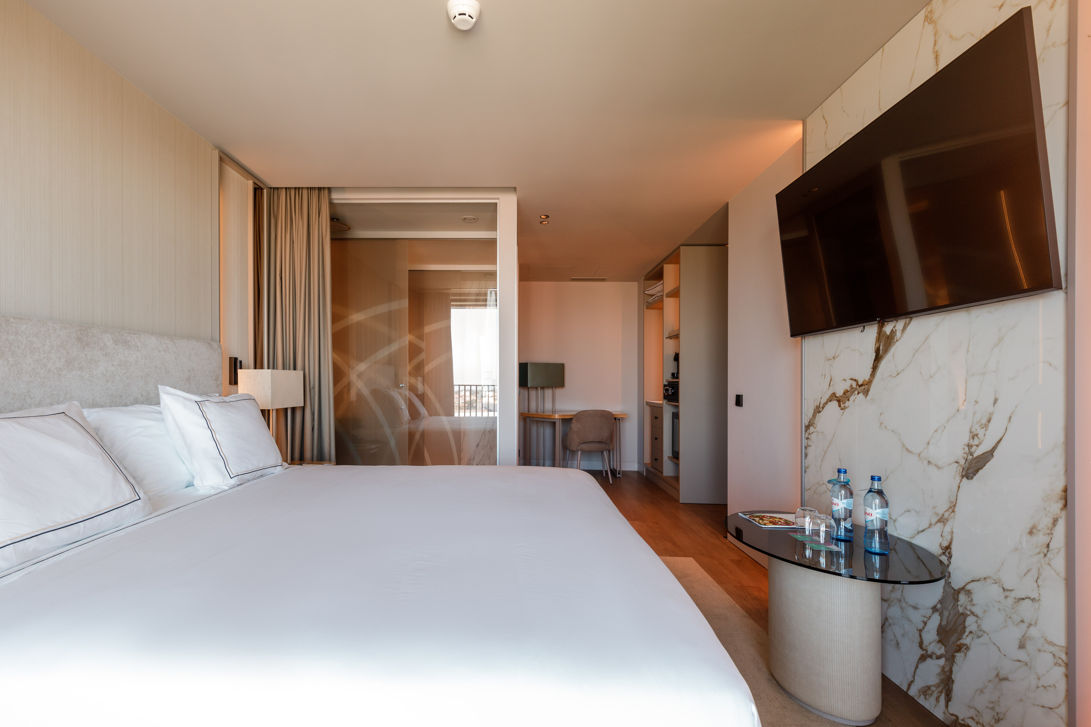 Modern hotel suite with king bed, smart TV, marble wall, and glass partition.