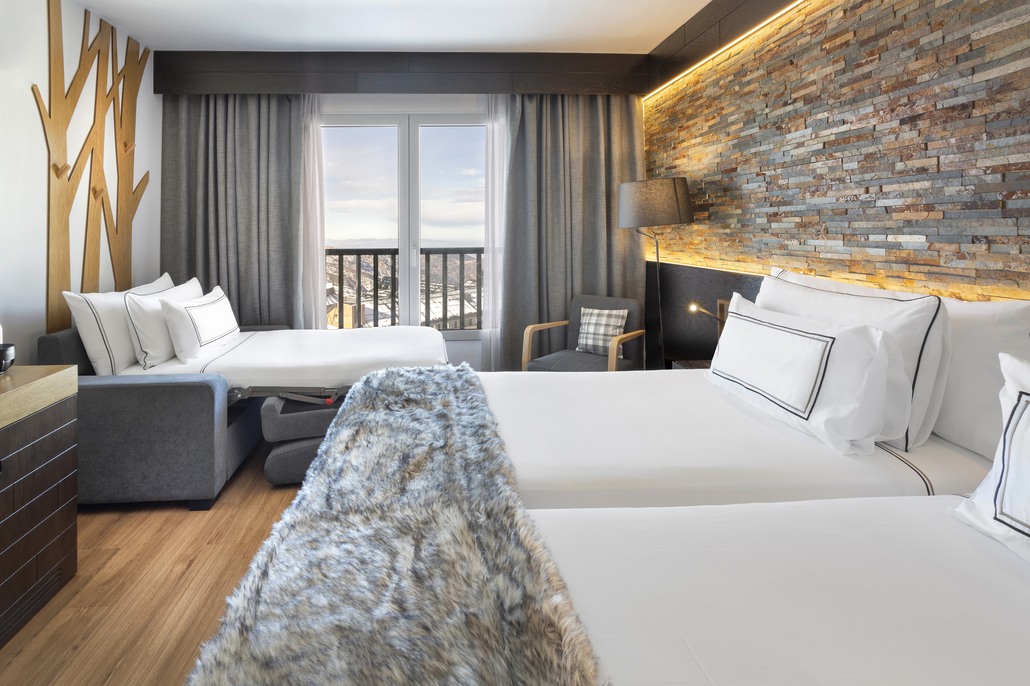 Modern ski resort room with sofa bed, stone wall, and mountain view.