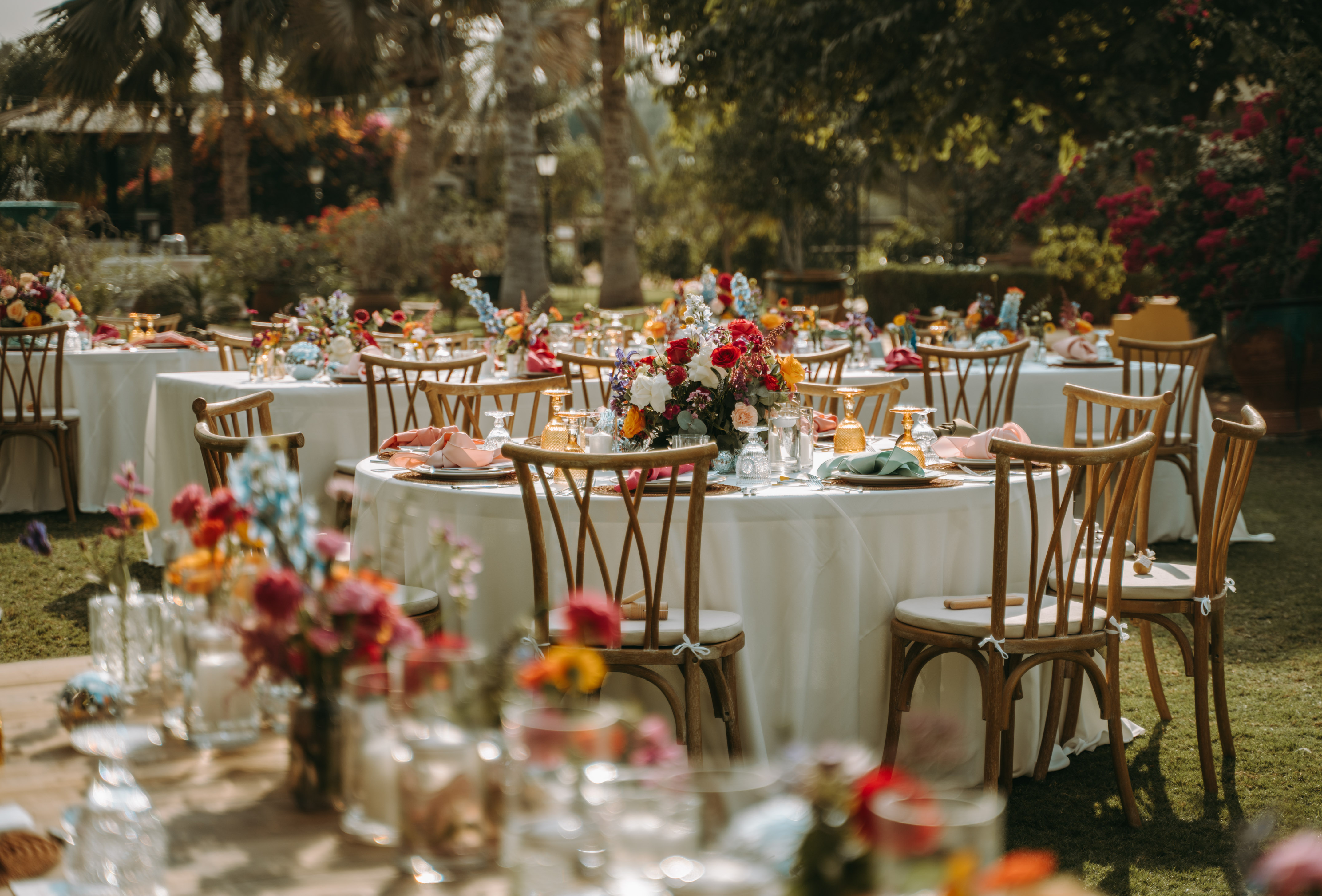 Outdoor tables with colorful floral centerpieces and wooden chairs in a lush garden setting.