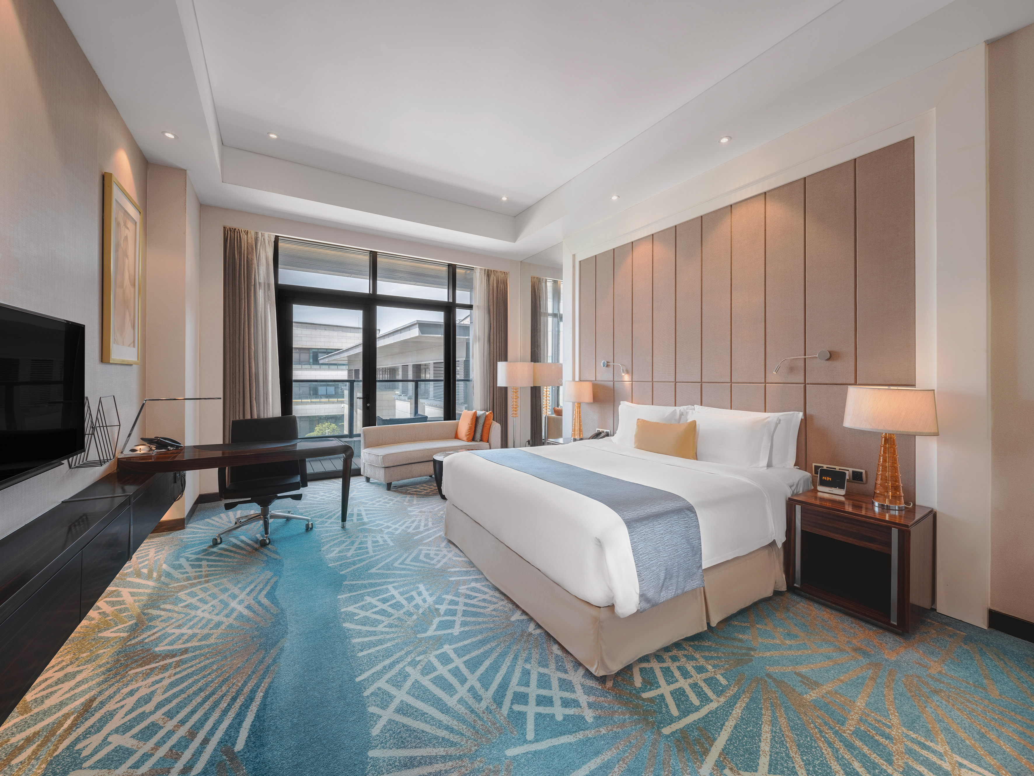 Modern hotel room with king bed, blue patterned carpet, desk, city view.