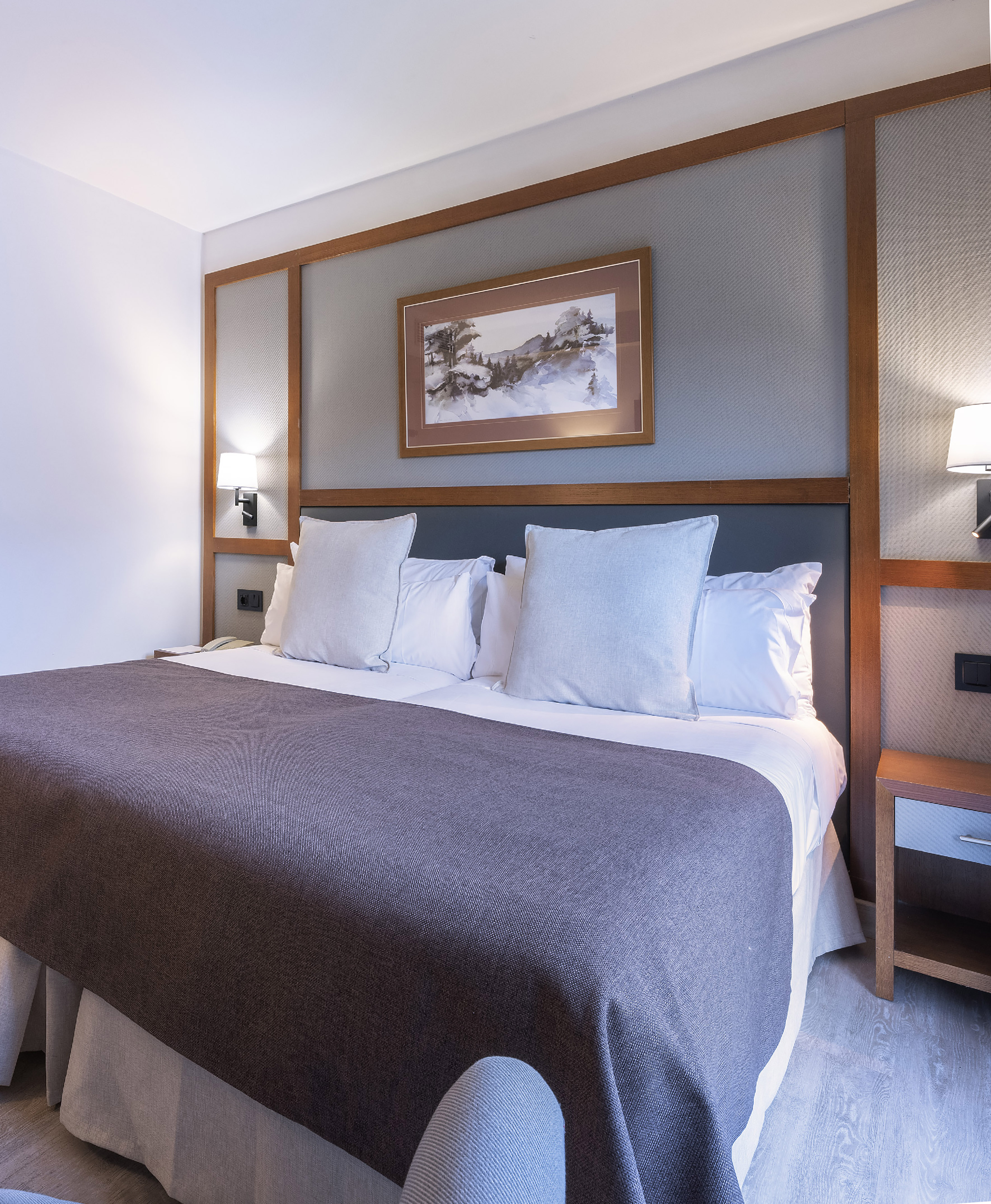 Modern hotel room with king bed, grey headboard, landscape art, and bedside lamps.