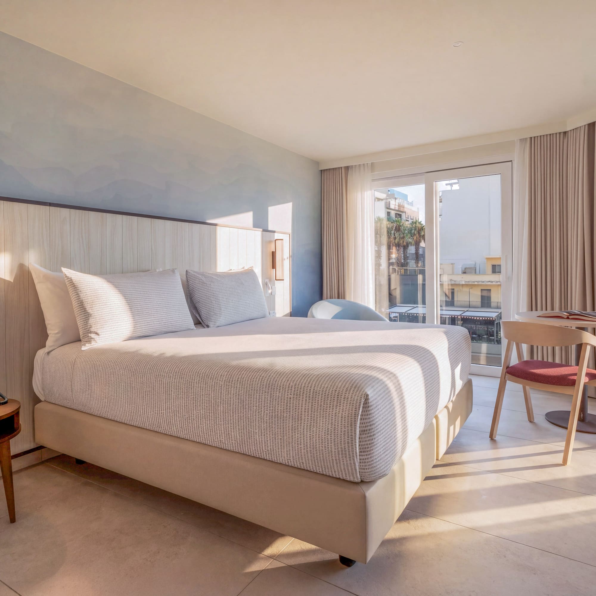 Sunlit modern hotel room with large bed, wooden headboard, and balcony view.