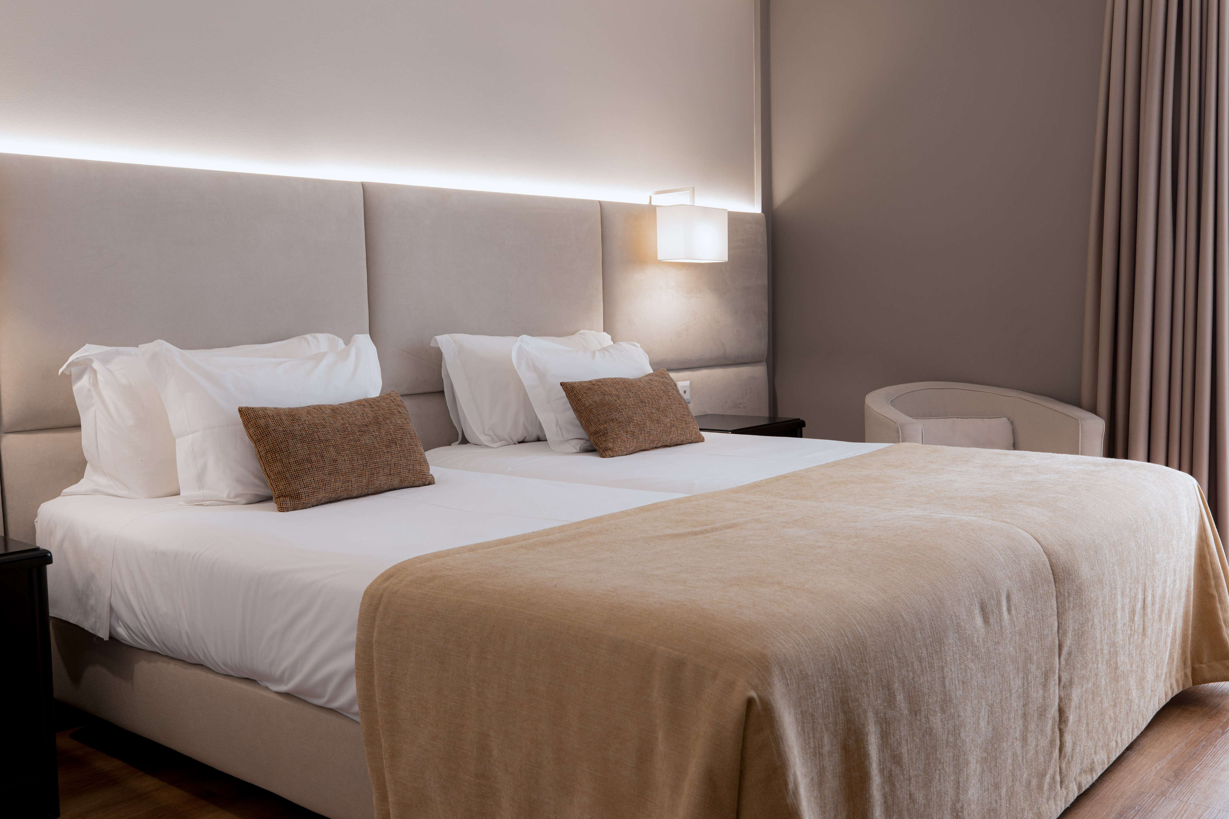 Modern hotel room with two beds, upholstered headboard, bedside lamp, and armchair.