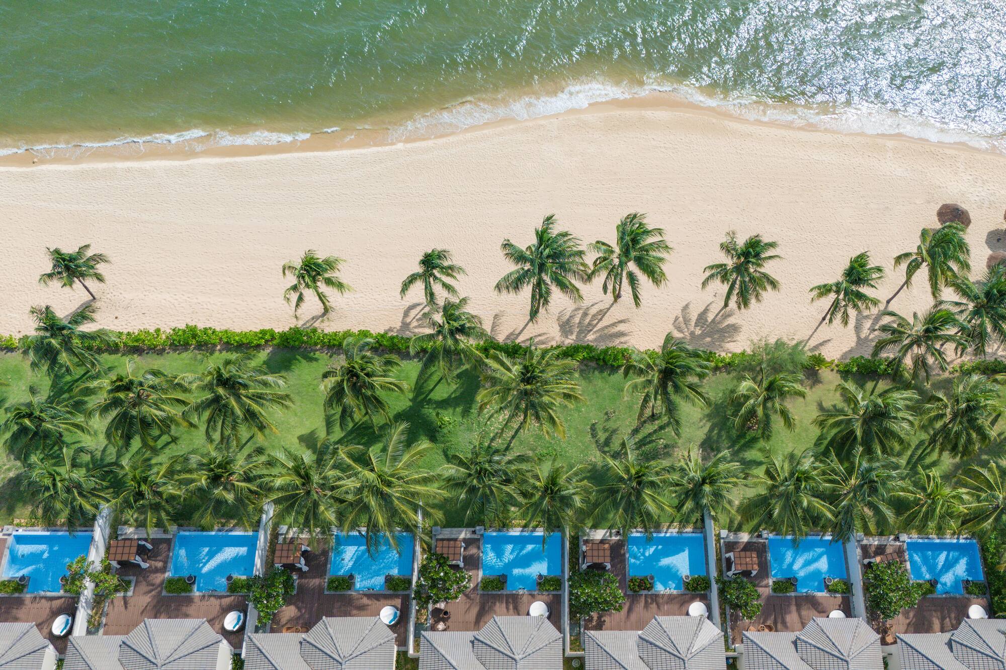Tropical beach resort aerial: ocean, palm trees, villas with private pools.