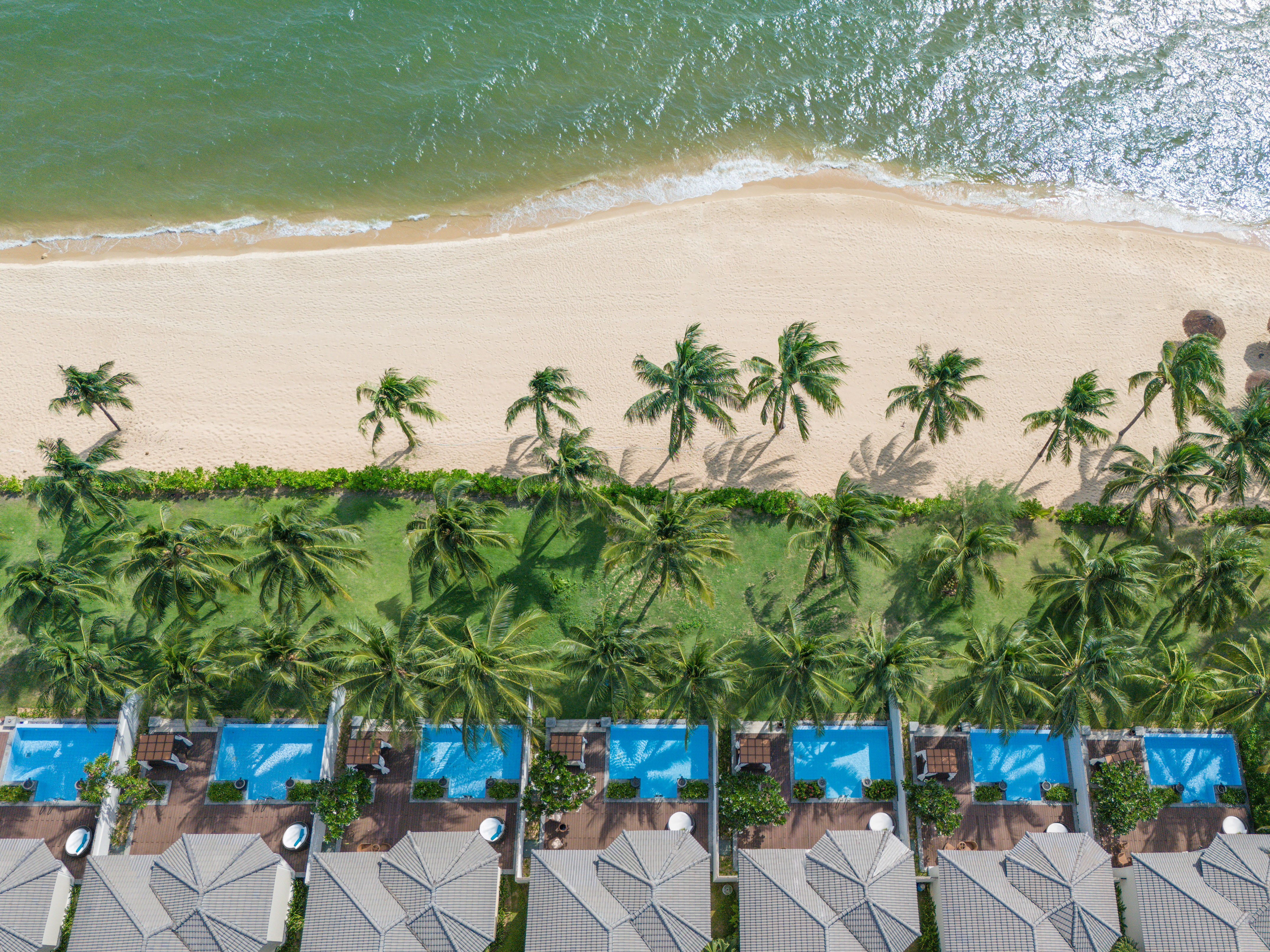 Tropical beach resort aerial: ocean, palm trees, villas with private pools.