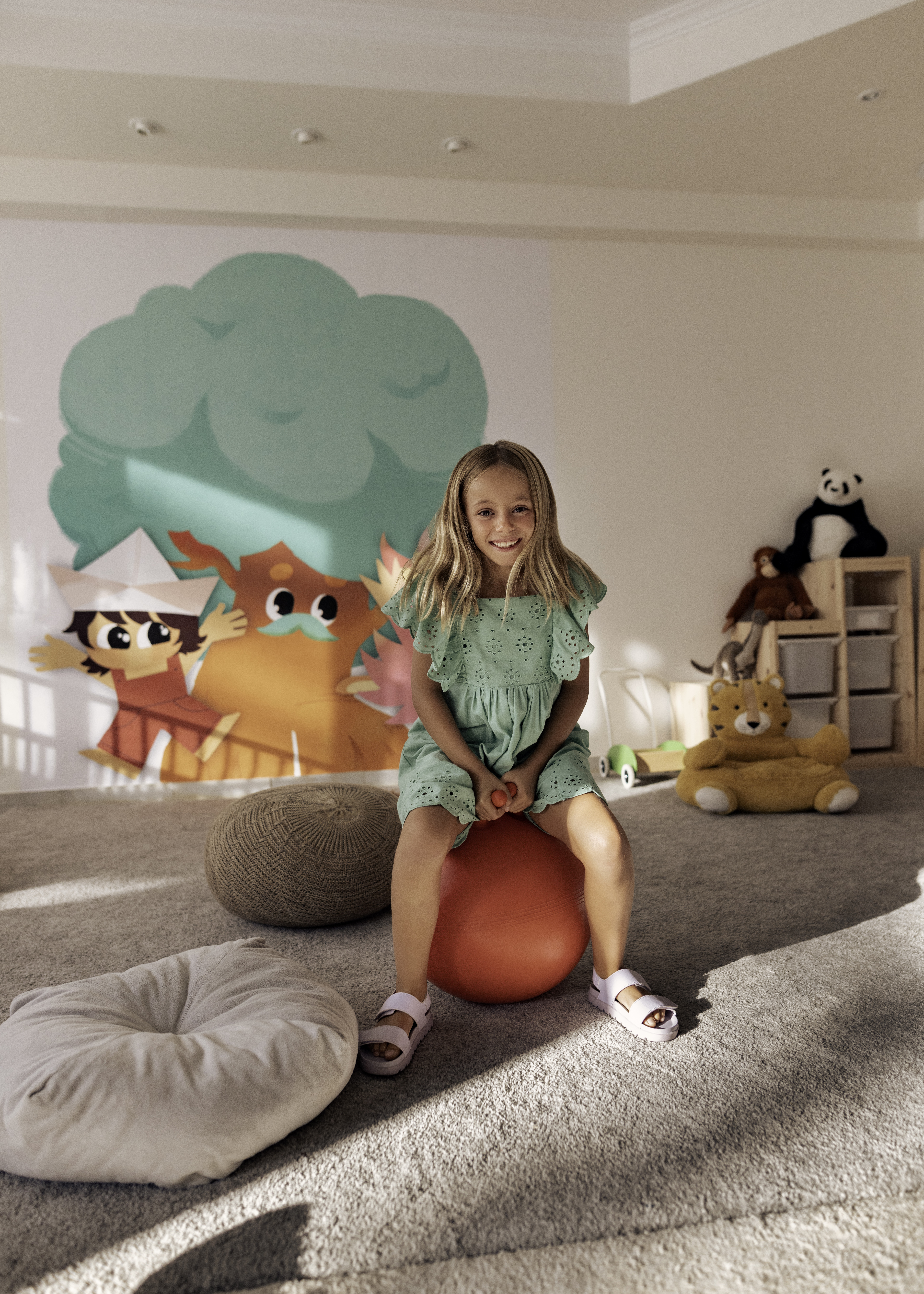 Young girl smiling on red hopper ball in playroom with mural and toys.