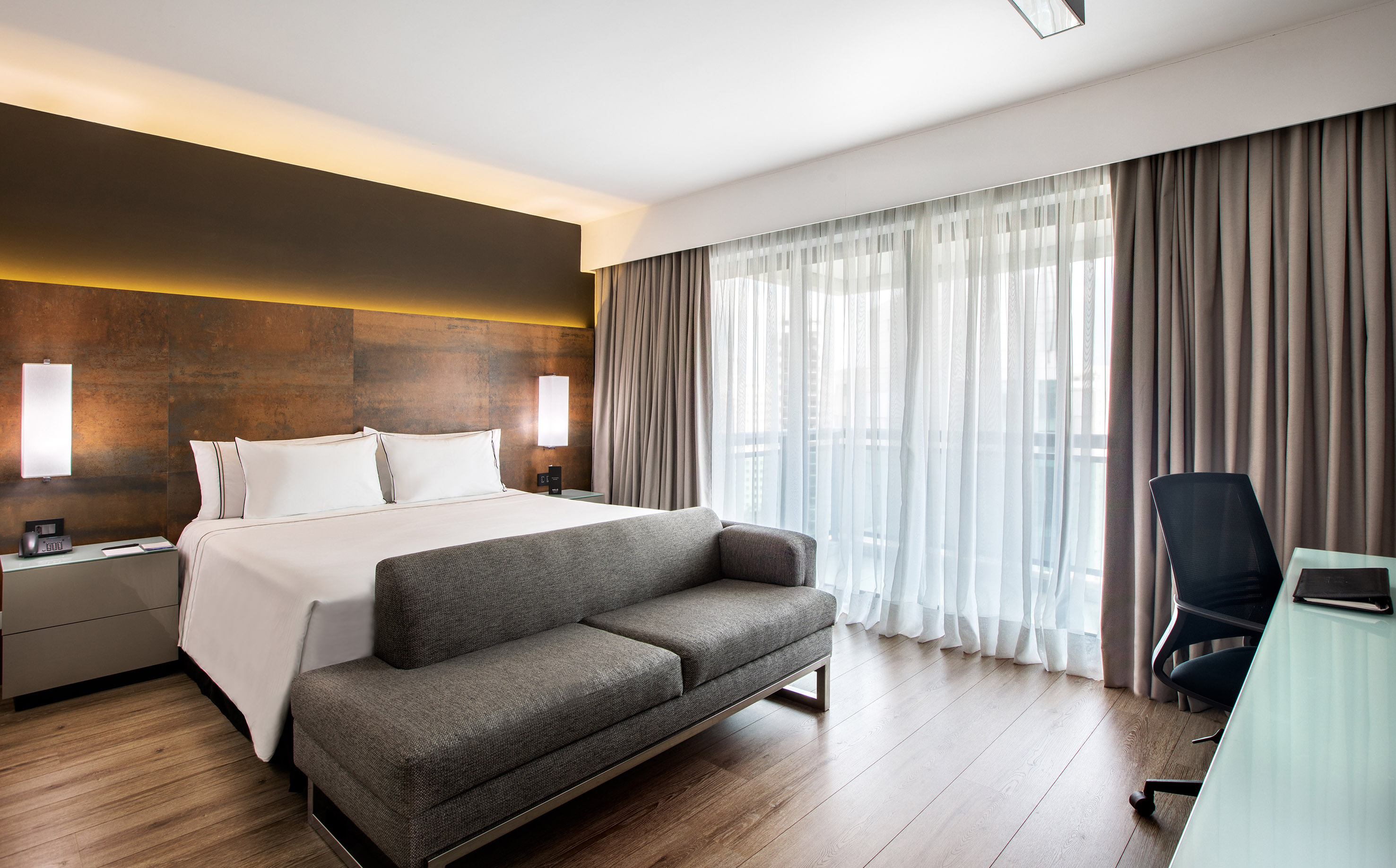 Contemporary hotel room with king bed, sofa, desk, and bright window.