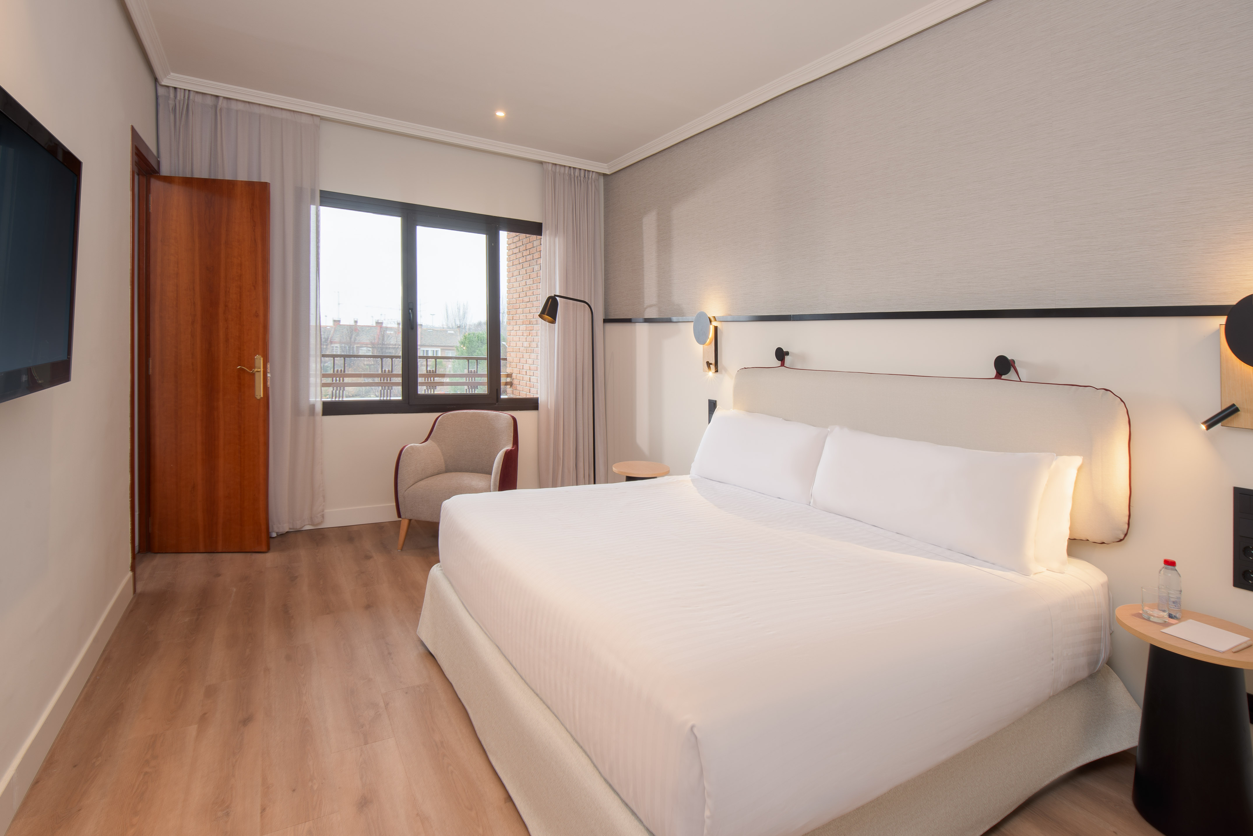 Modern hotel bedroom with white king bed, wooden floor, window, and armchair.