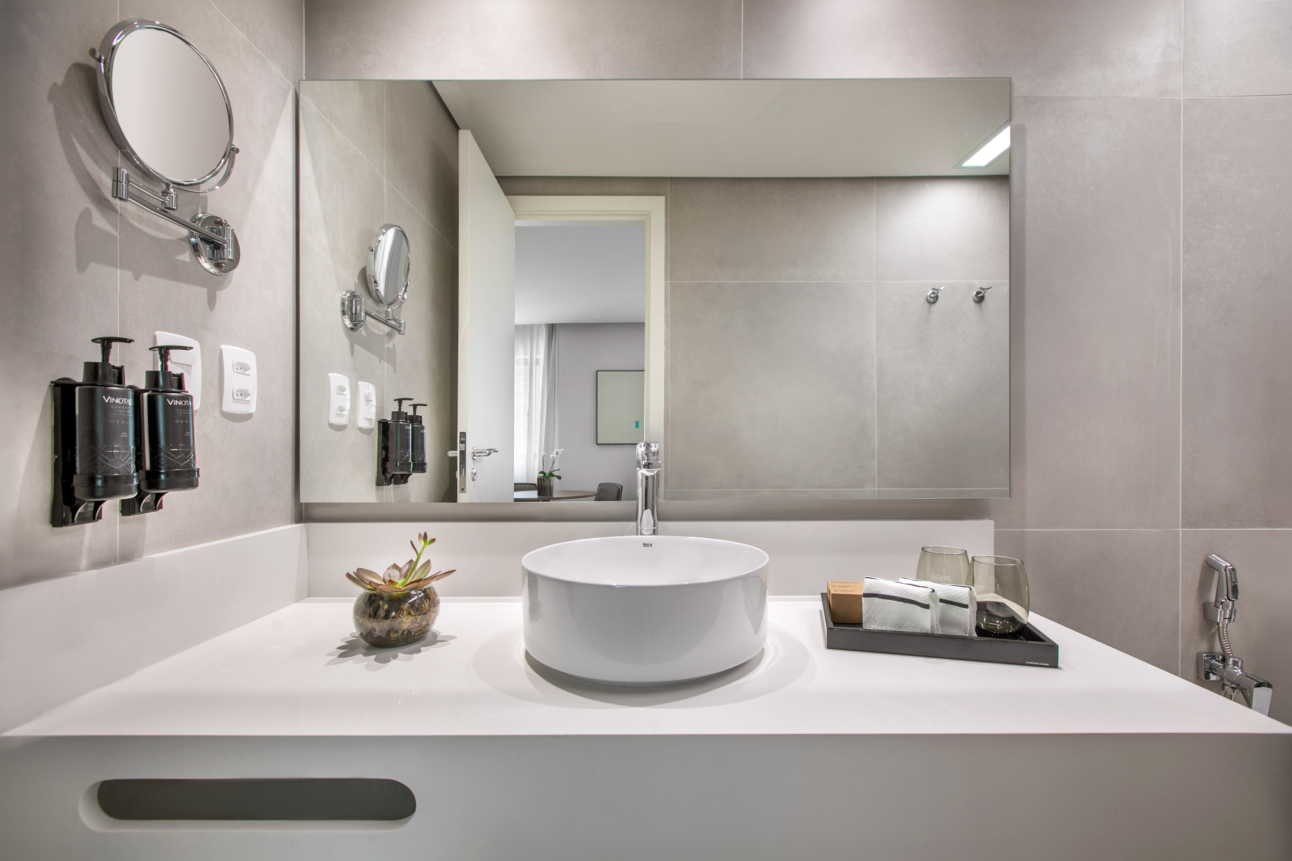 Modern hotel bathroom vanity with white vessel sink, large mirror, and grey tiled walls.