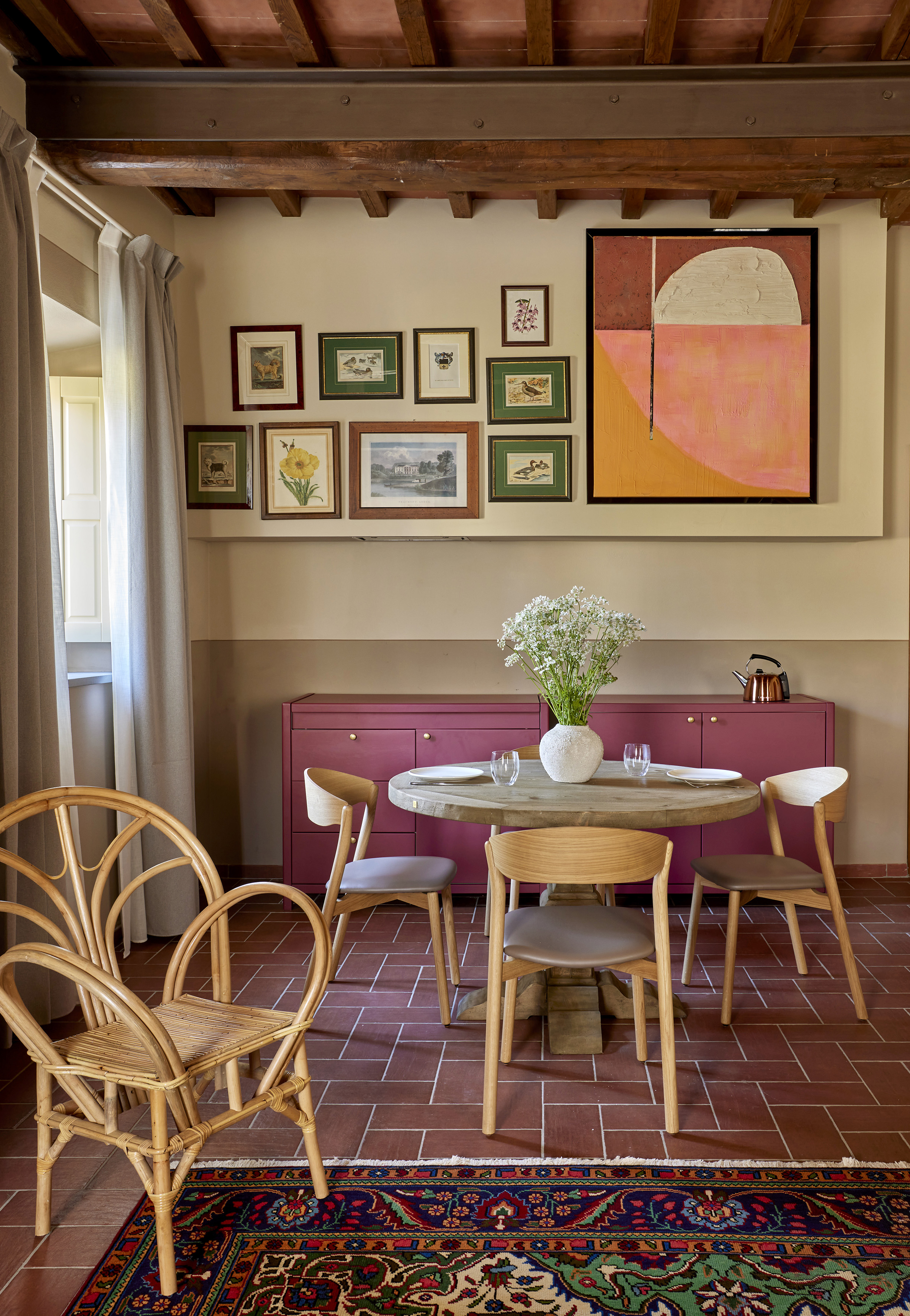 Dining area with magenta cabinet, gallery wall, wood table, terracotta floor.