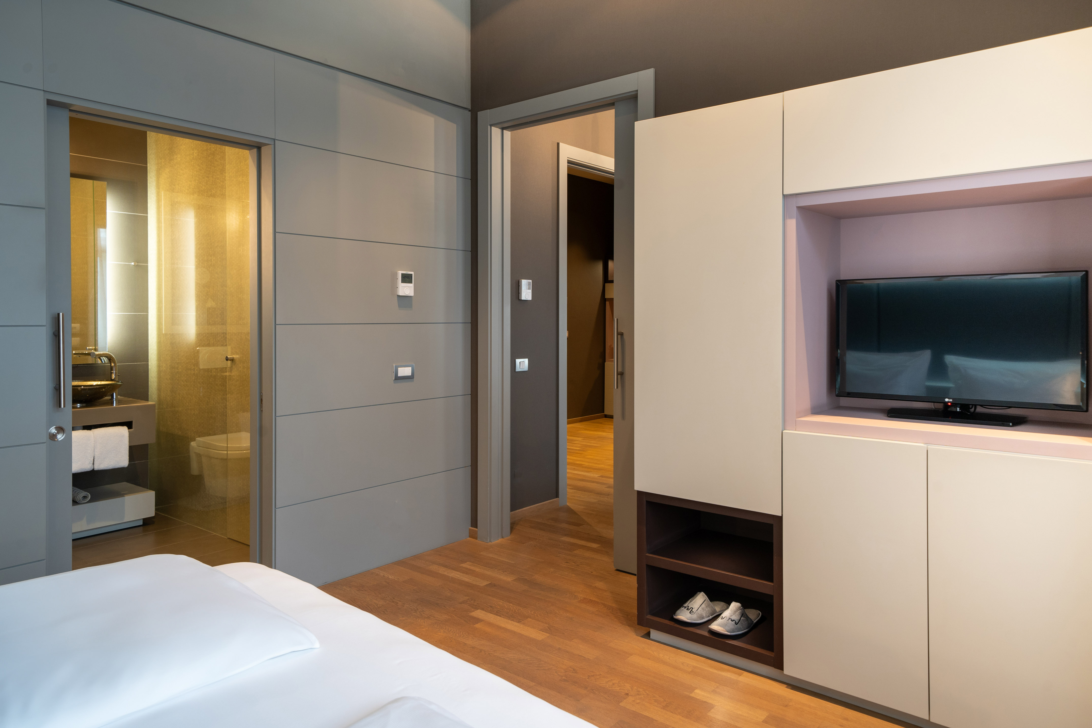 Contemporary hotel bedroom with TV, wooden floor, grey walls, and visible bathroom.