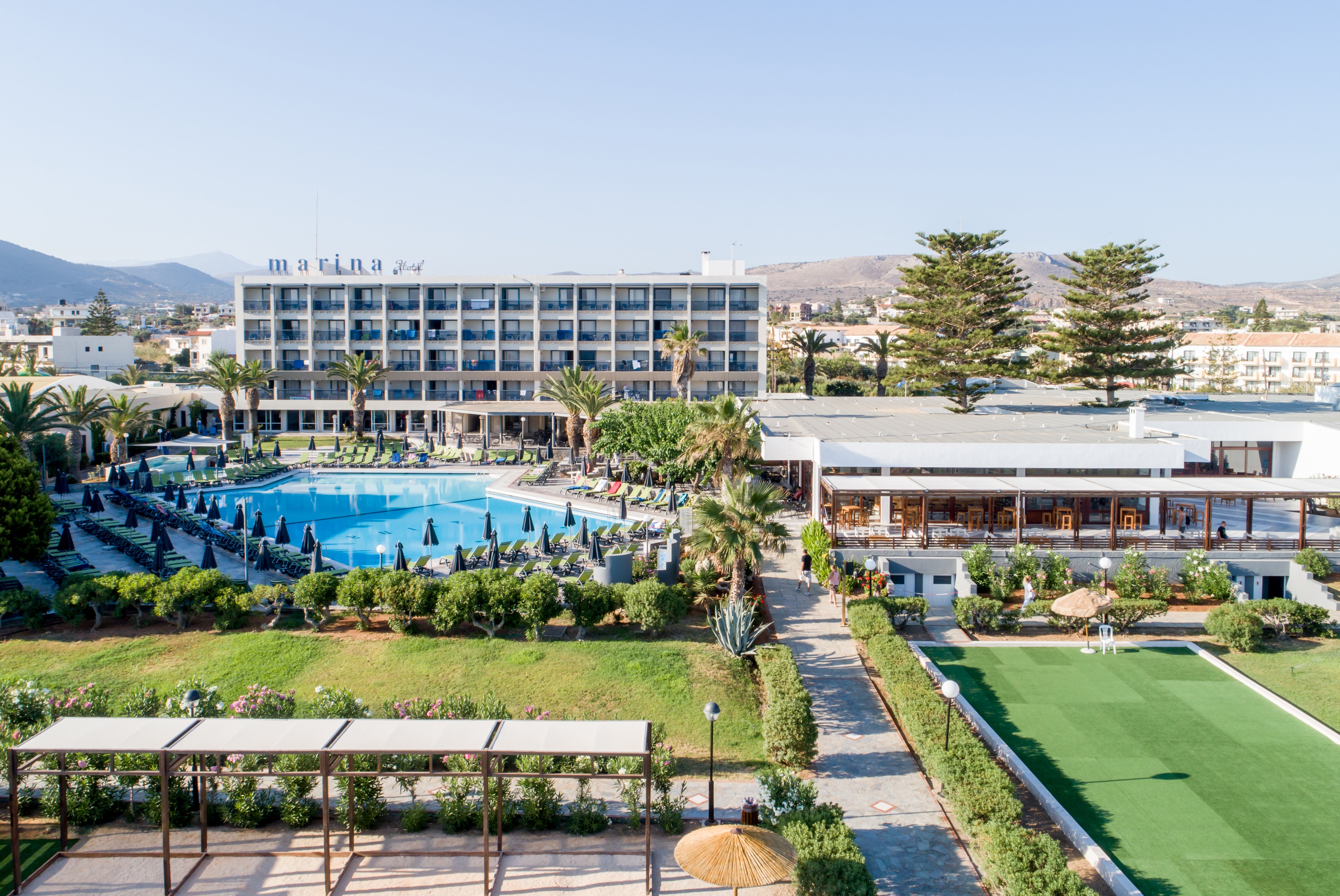 Marina Hotel resort with pool, palm trees, green lawns, and mountain background.