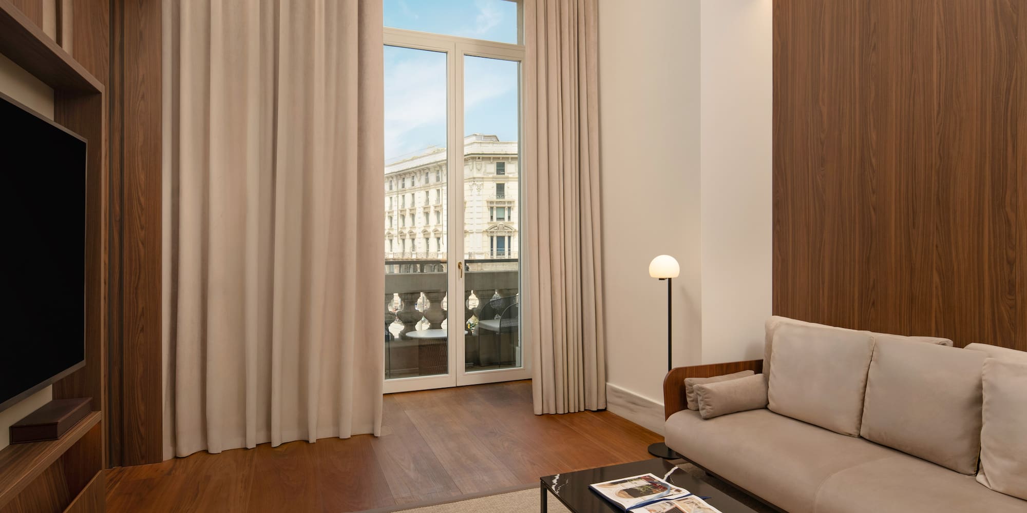 Modern hotel suite living area with city view balcony, sofa, hardwood floor.