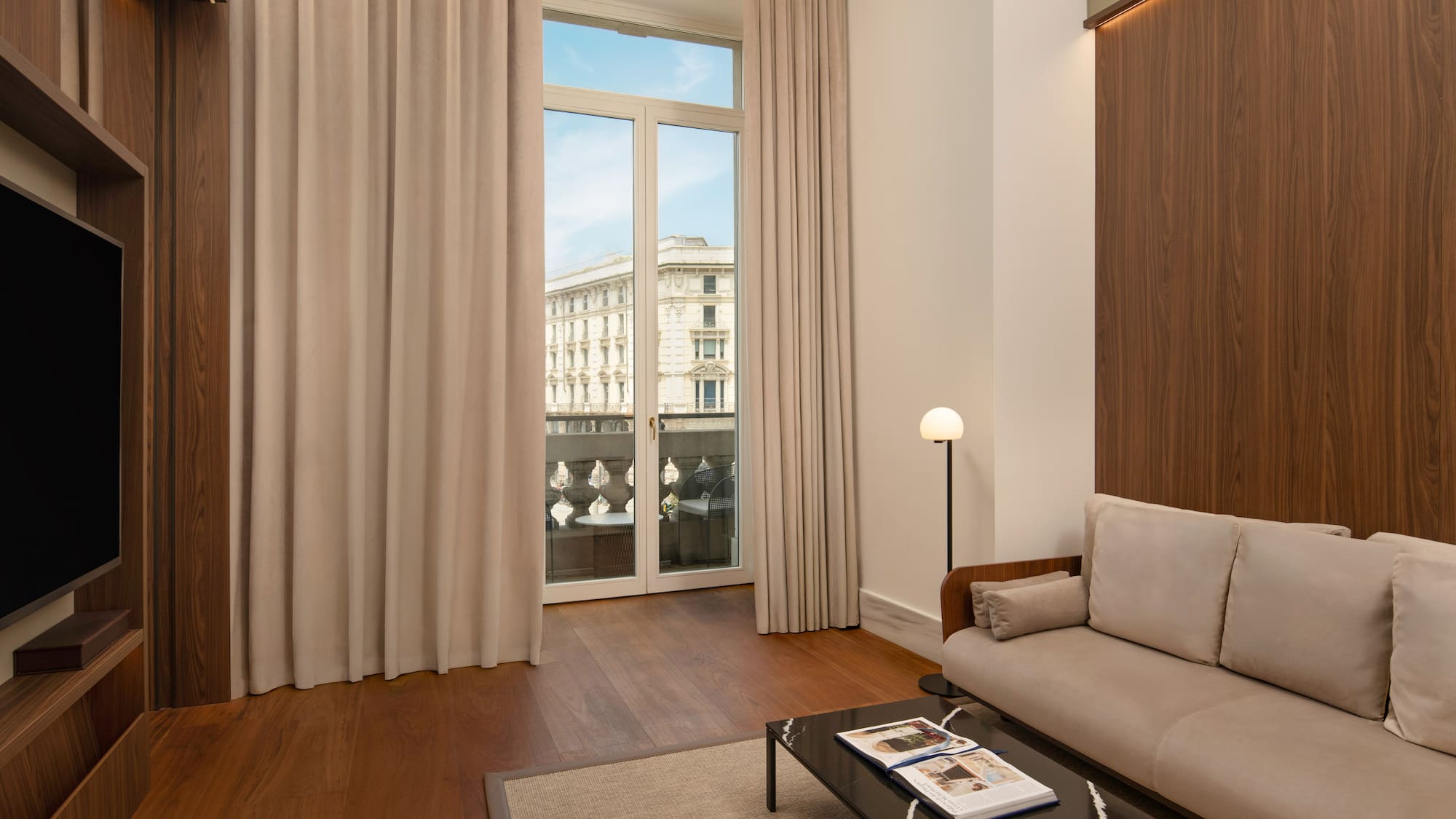 Modern hotel suite living area with city view balcony, sofa, hardwood floor.