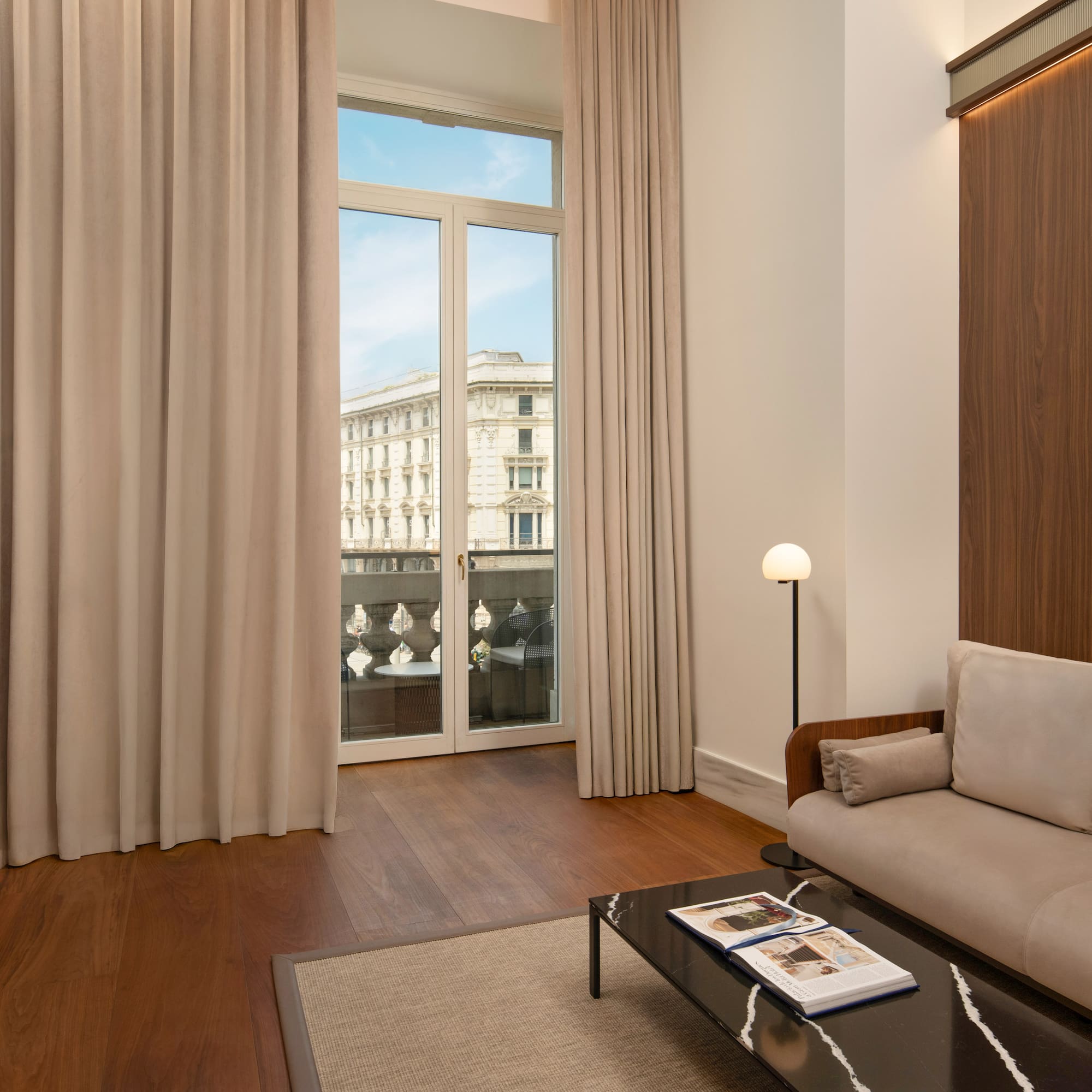 Modern hotel suite living area with city view balcony, sofa, hardwood floor.