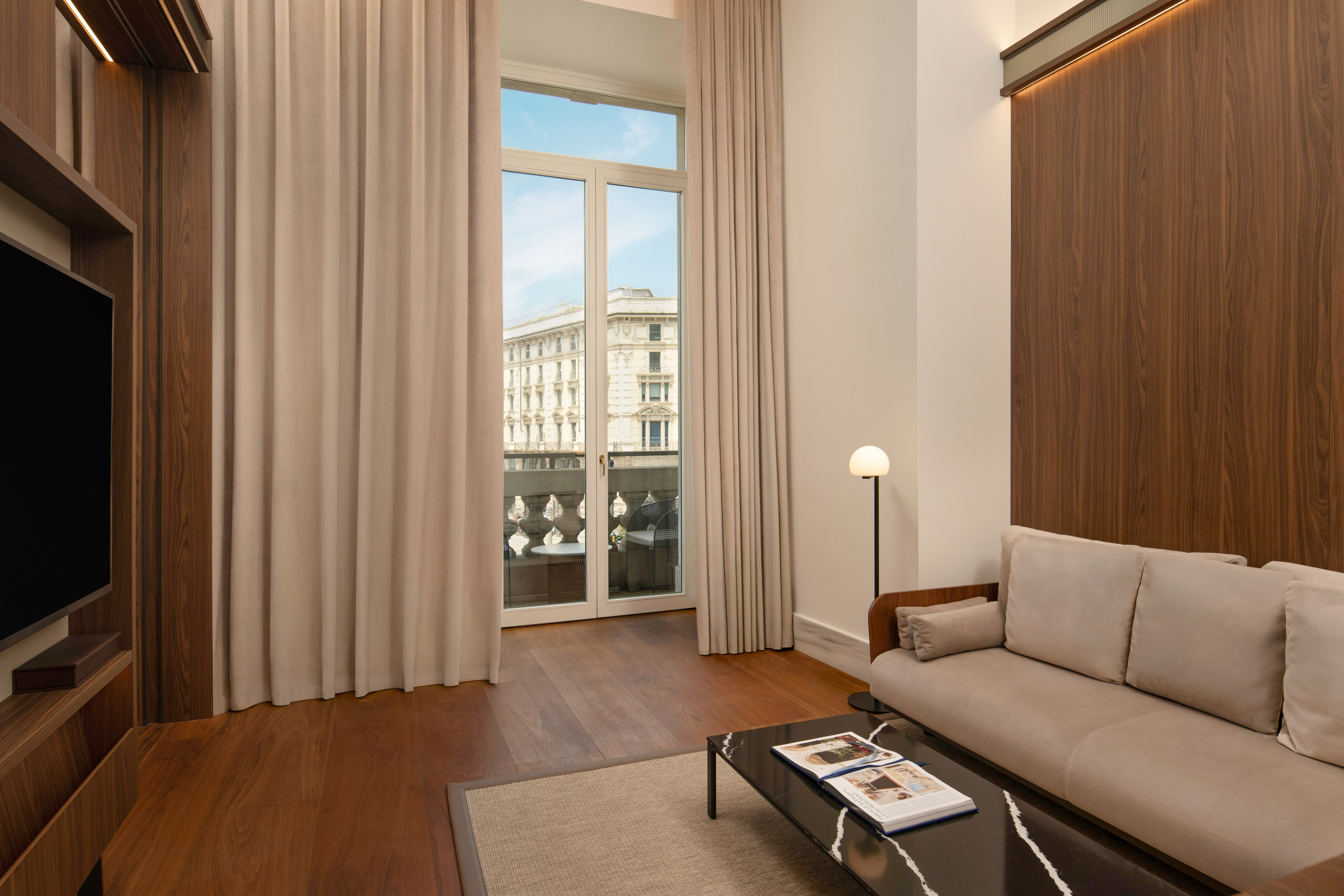 Modern hotel suite living area with city view balcony, sofa, hardwood floor.