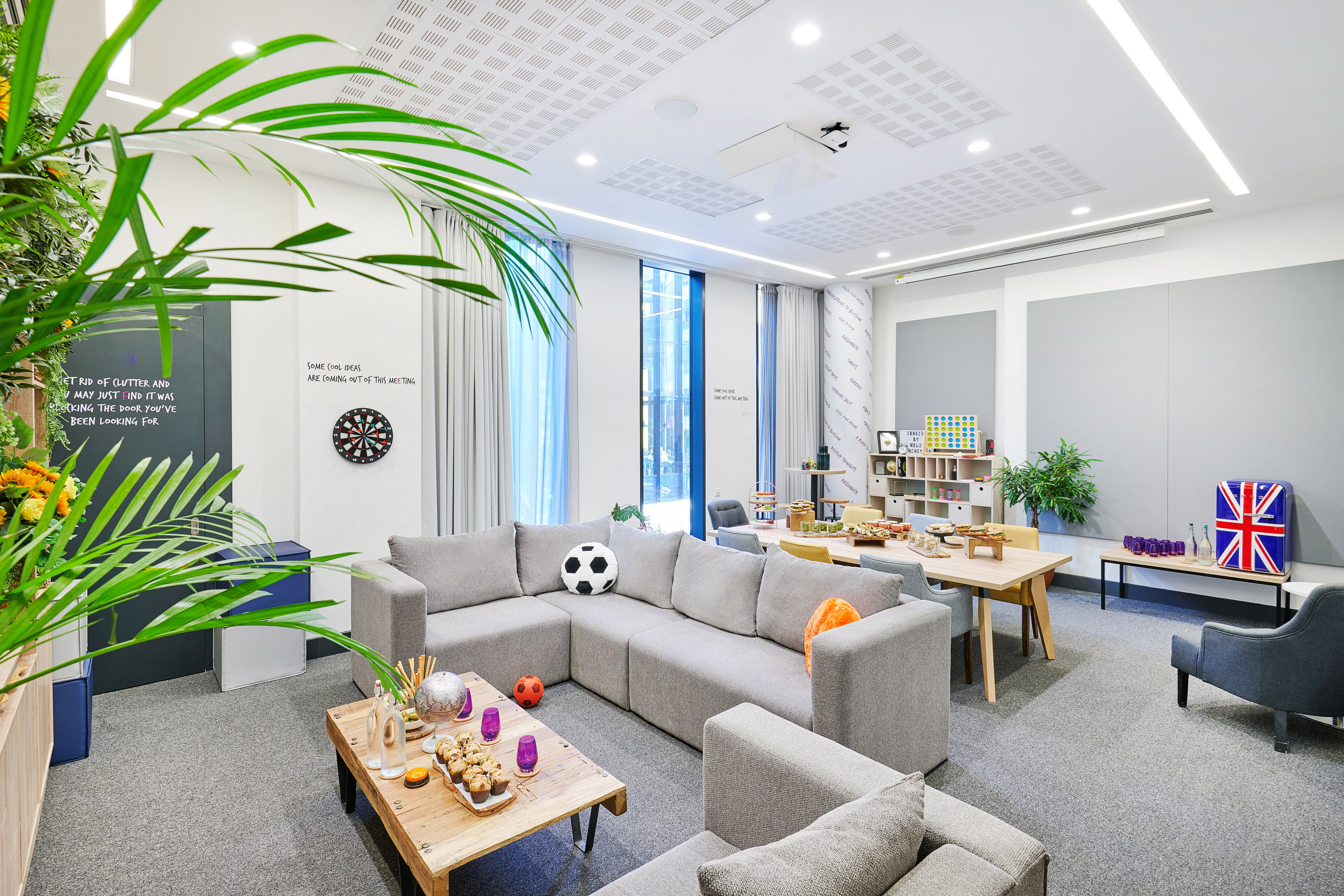 Modern office lounge area with grey sofa, dartboard, snacks, and communal table.