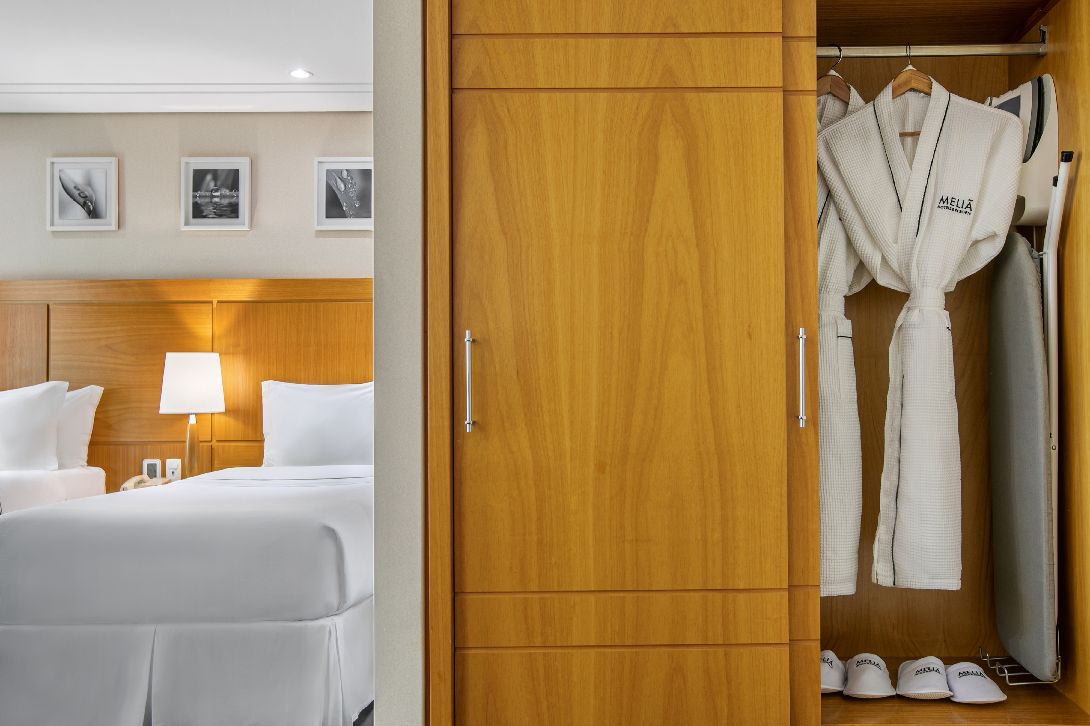Hotel bedroom with white bed, wooden closet, Meliá bathrobes, and slippers.
