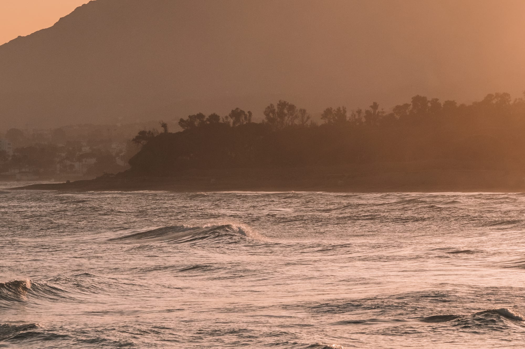 Atardecer en la costa con montañas, olas suaves y playa rocosa.