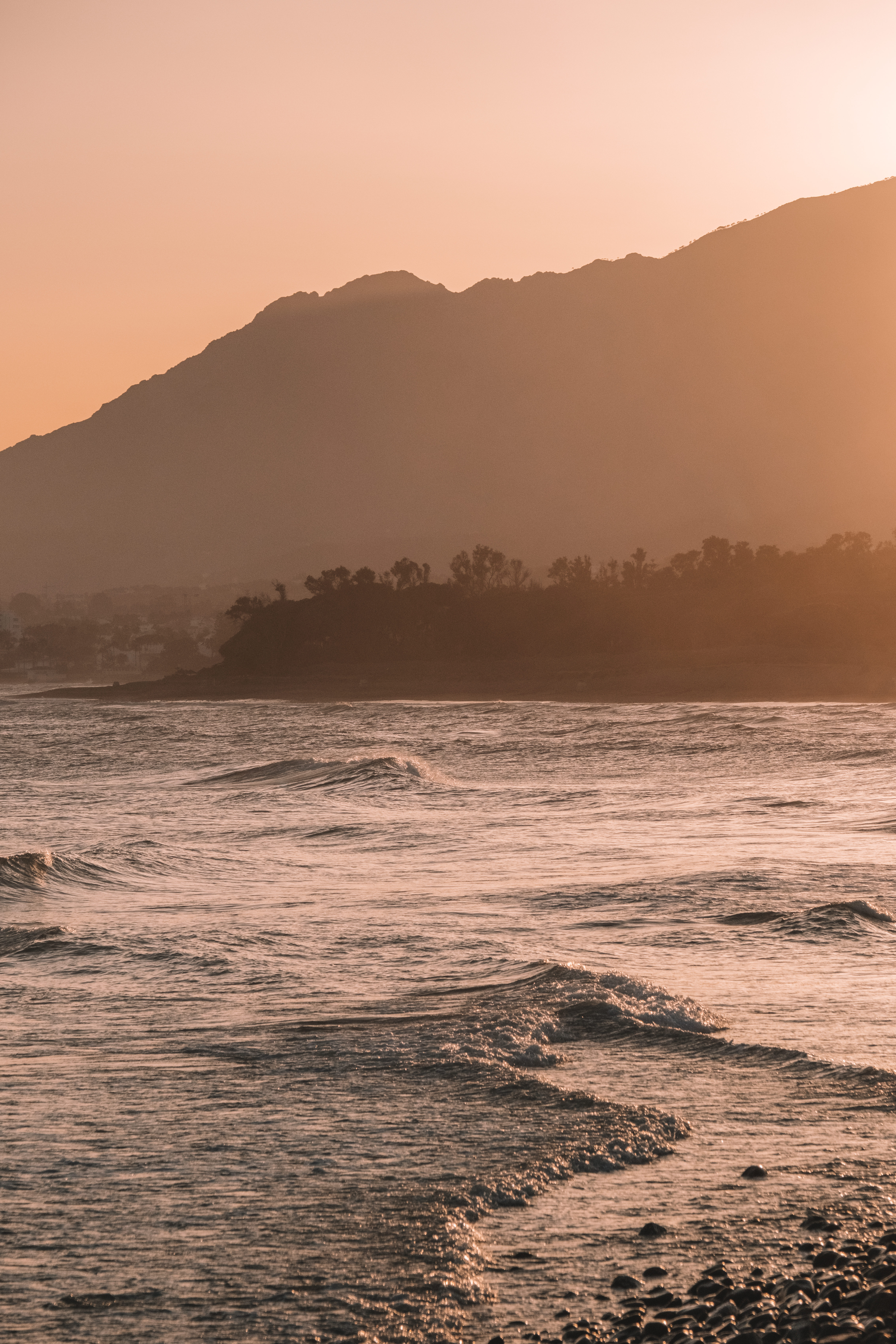 Atardecer en la costa con montañas, olas suaves y playa rocosa.