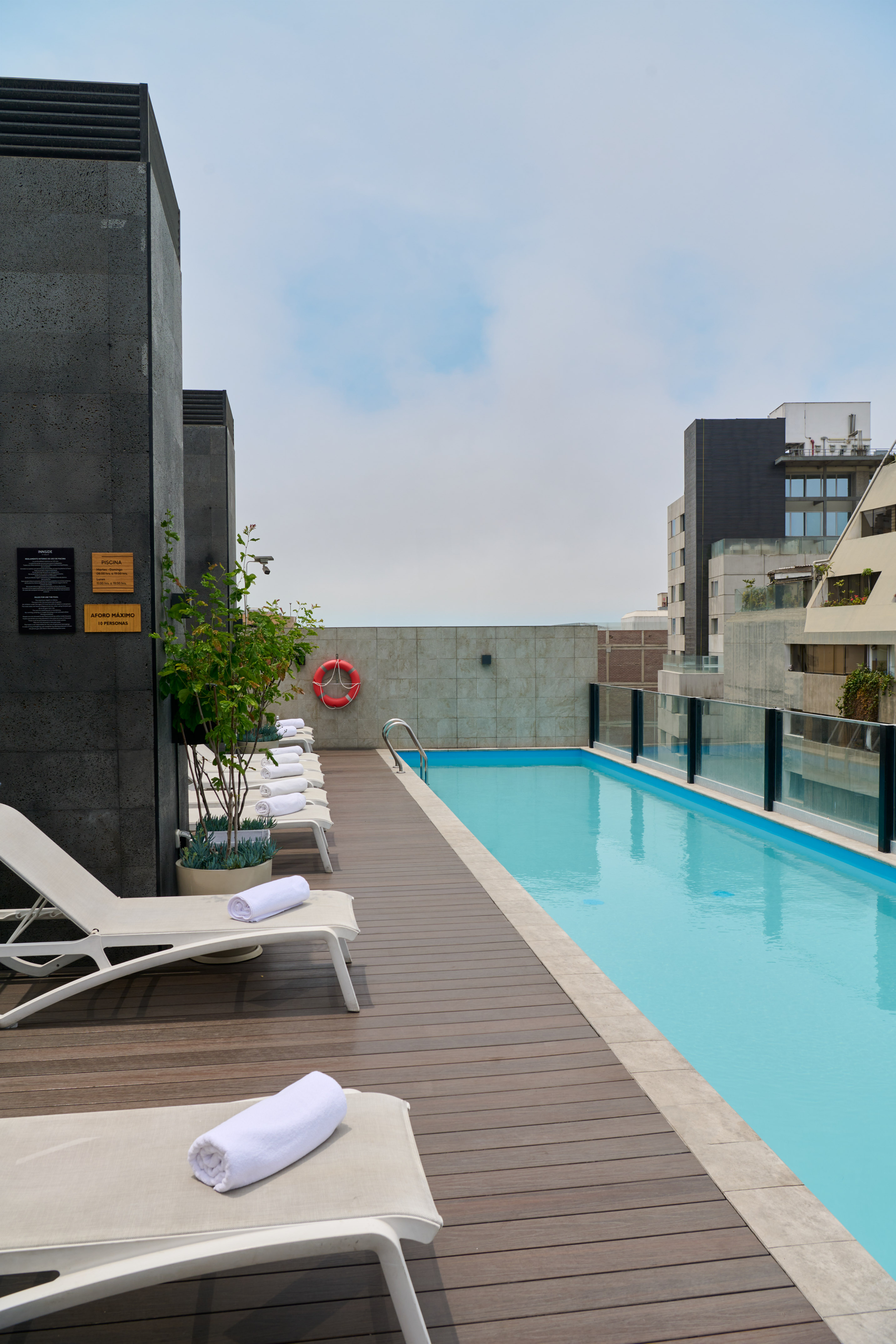 Rooftop swimming pool with wooden deck, lounge chairs, and urban buildings.