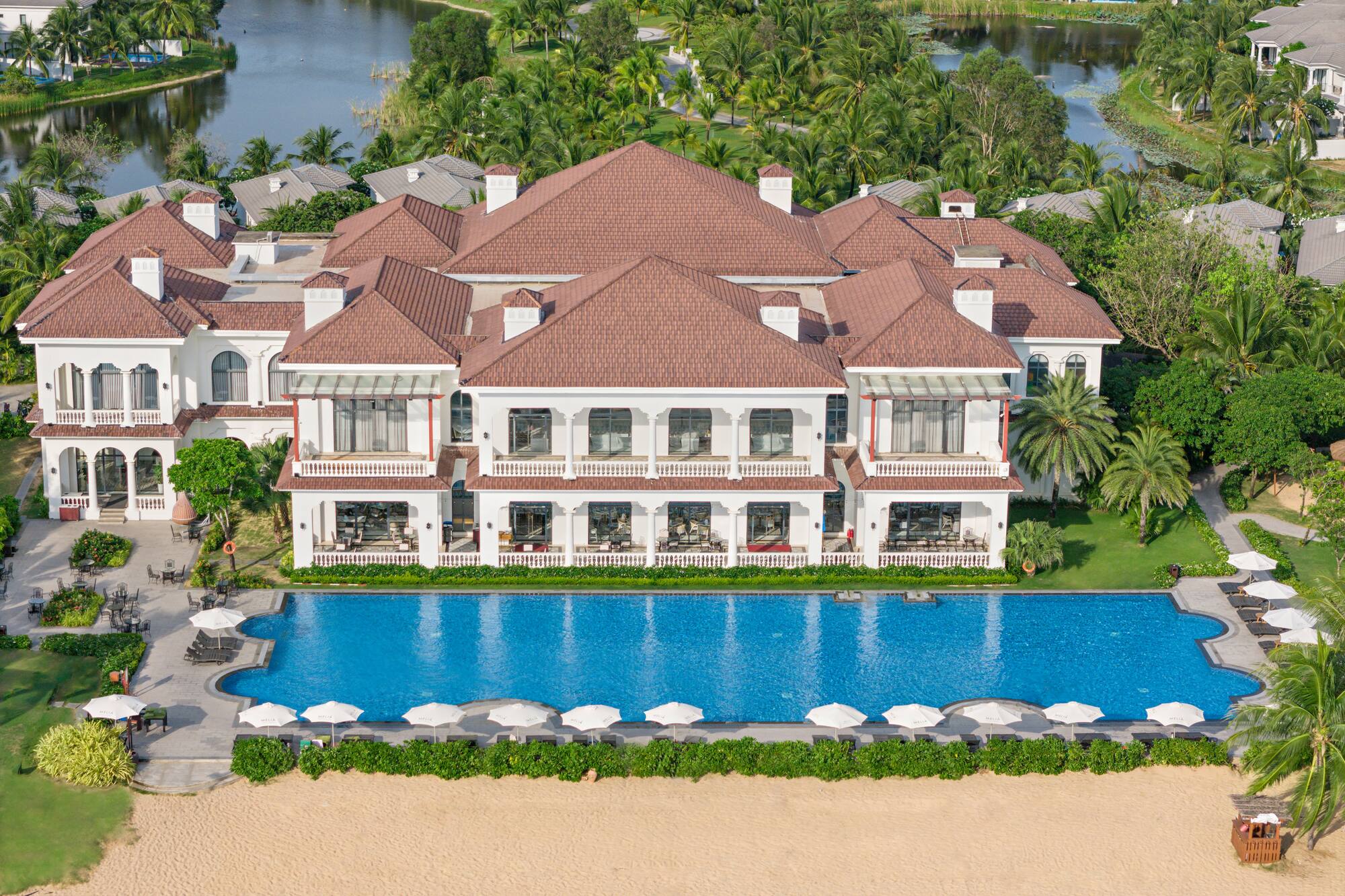 Luxury beachfront resort with large pool, white building, and tropical palm trees.