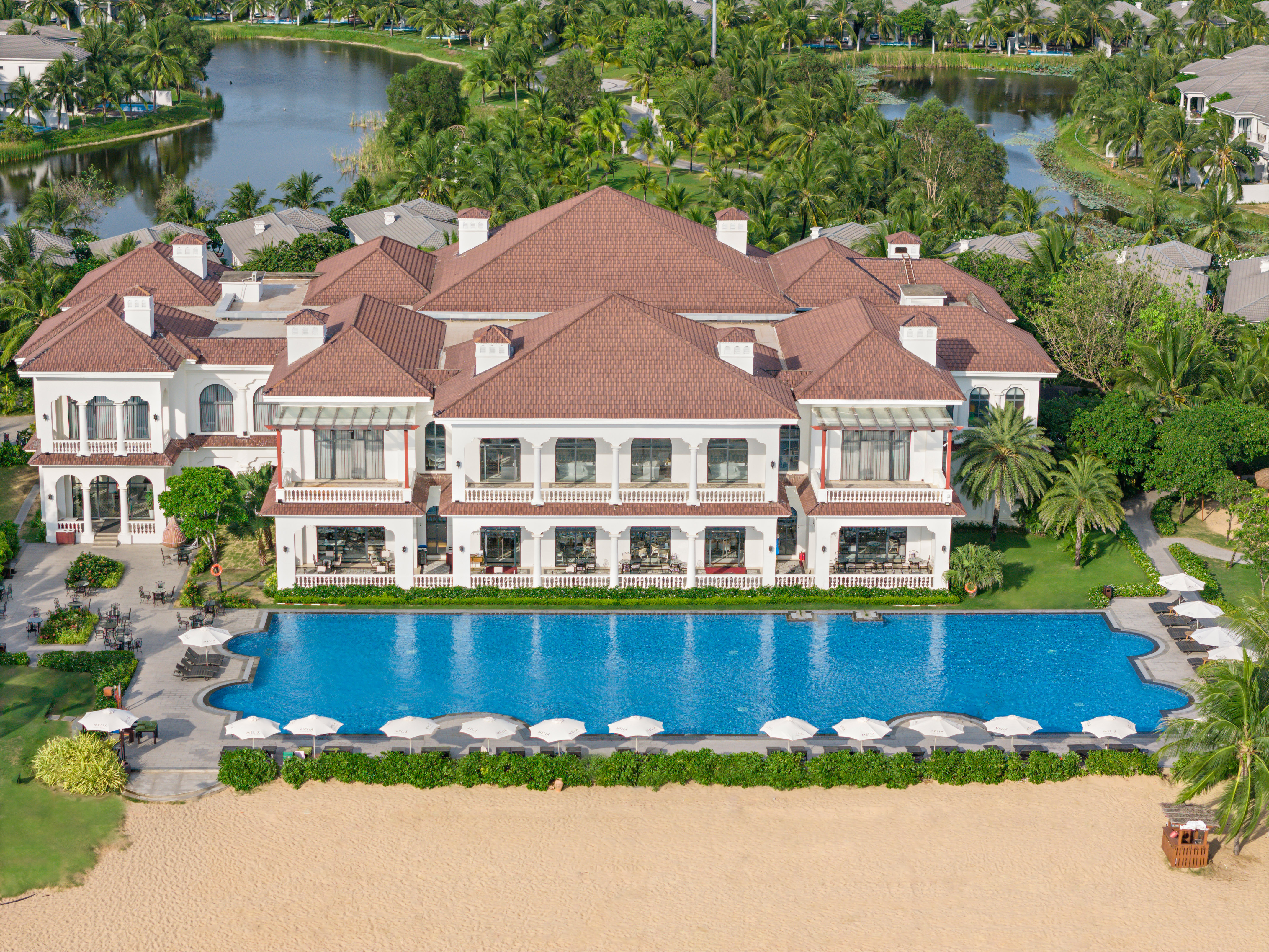 Luxury beachfront resort with large pool, white building, and tropical palm trees.