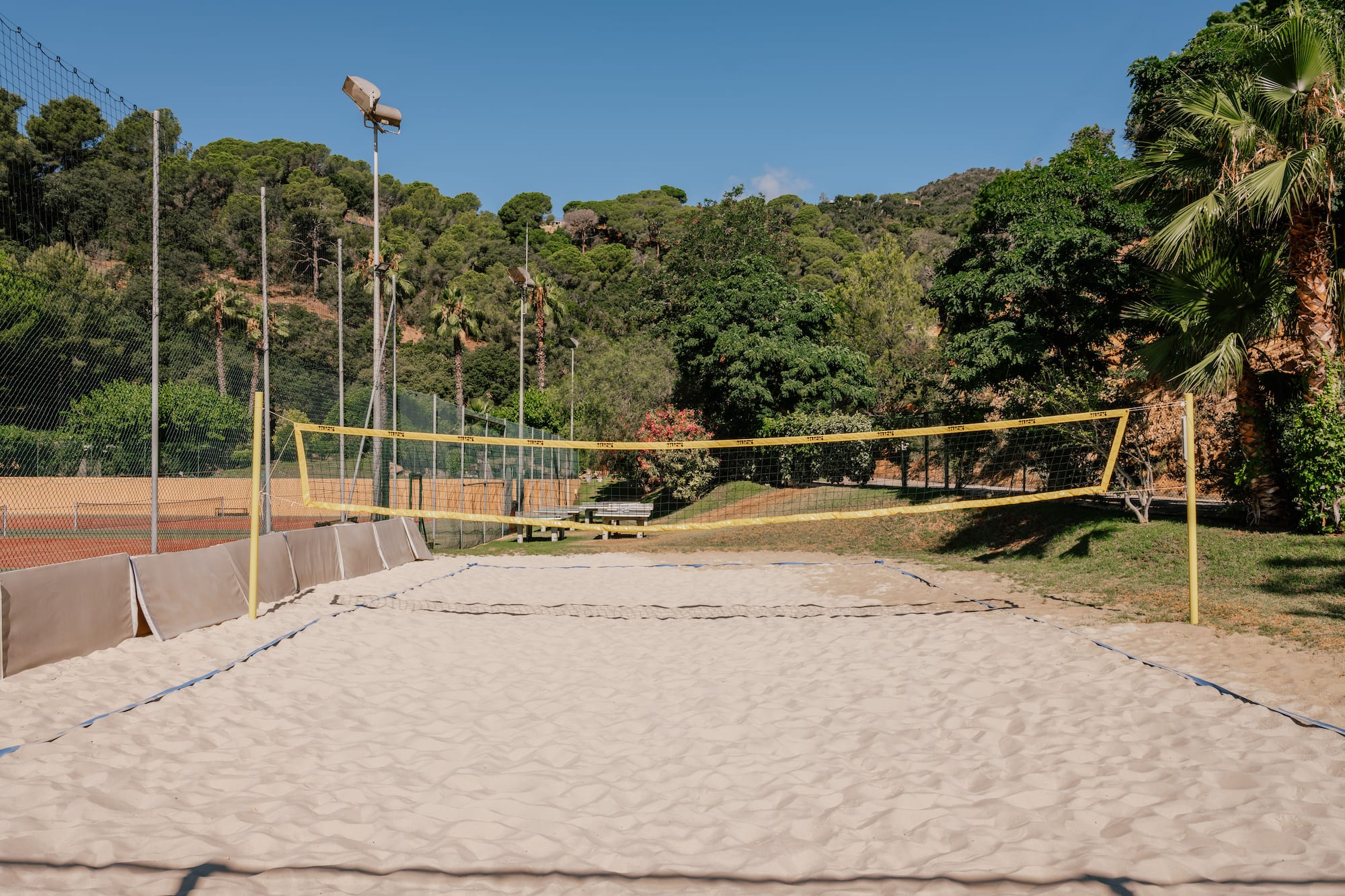 Sandy outdoor beach volleyball court with net, surrounded by green hills and palms.