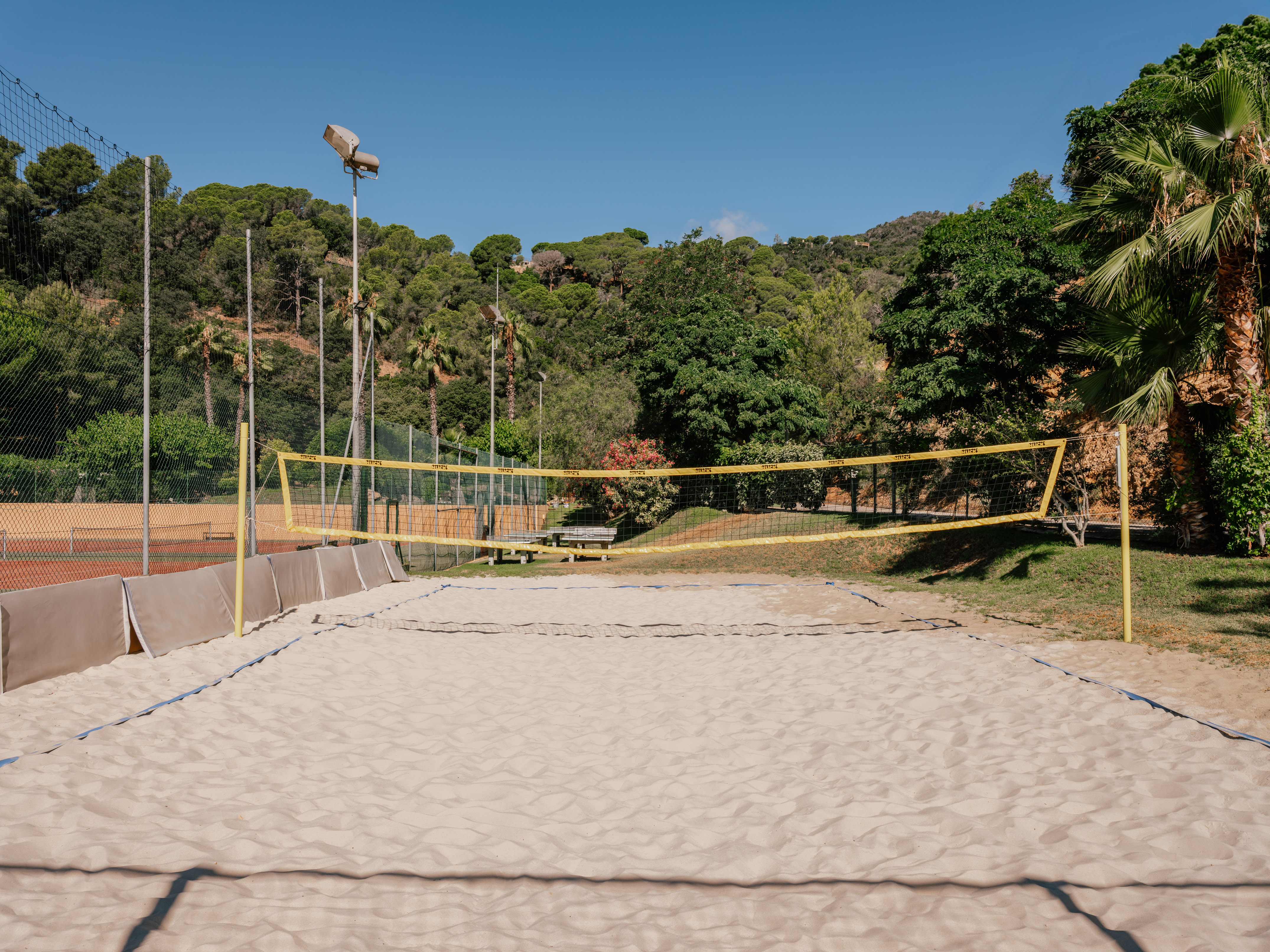 Sandy outdoor beach volleyball court with net, surrounded by green hills and palms.