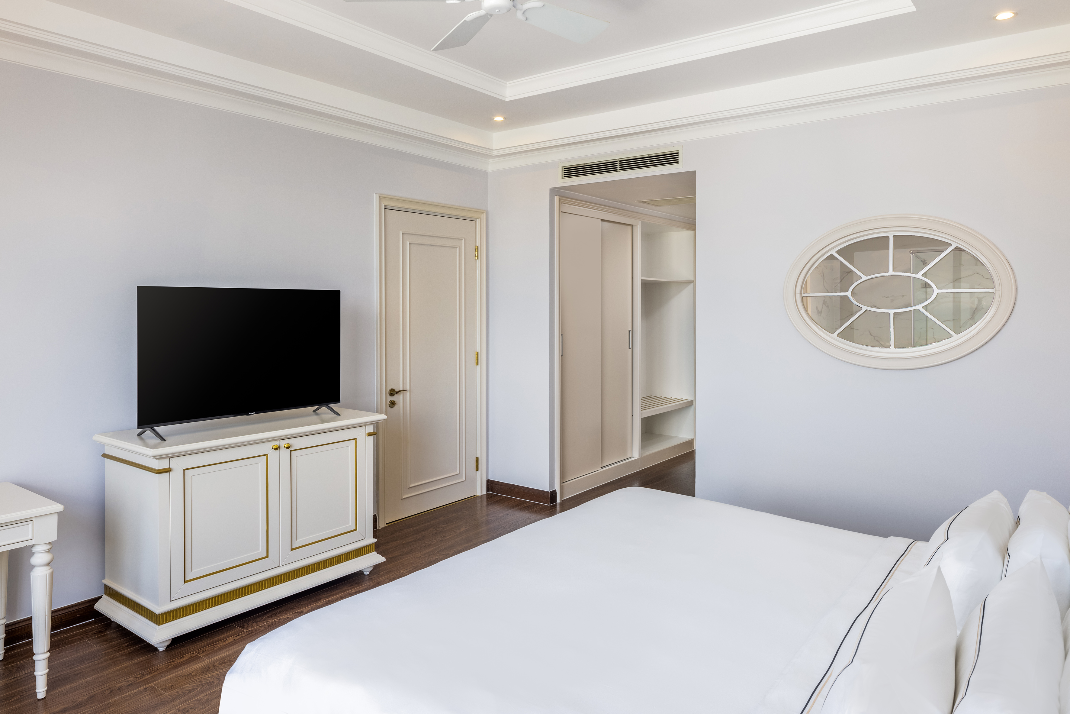 Elegant hotel room with white bed, TV, built-in closet, and oval mirror.