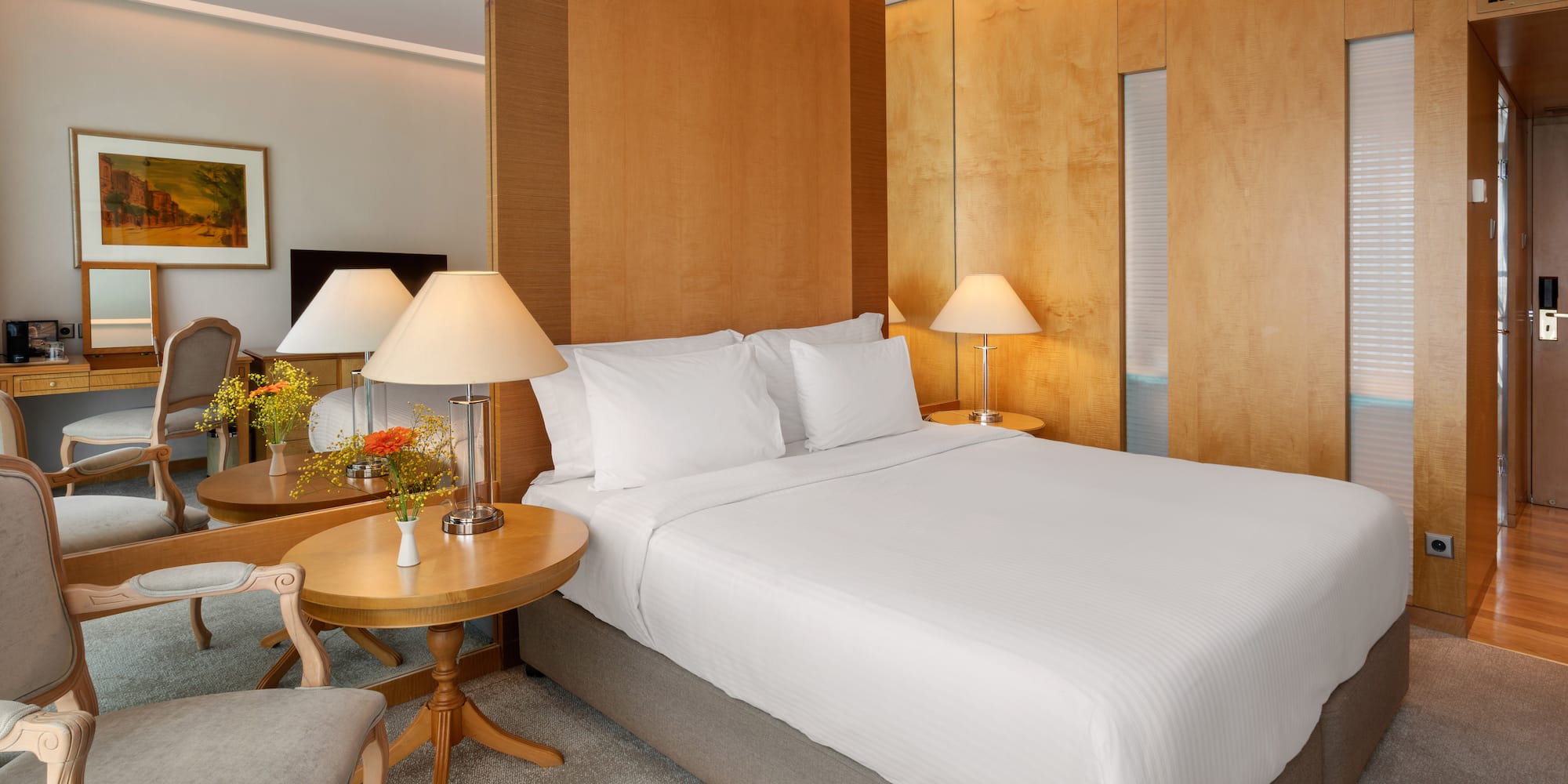 Elegant hotel bedroom with large bed, wooden panels, bedside lamps, and a vanity area.