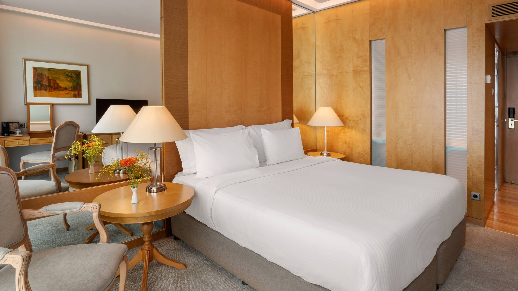 Elegant hotel bedroom with large bed, wooden panels, bedside lamps, and a vanity area.