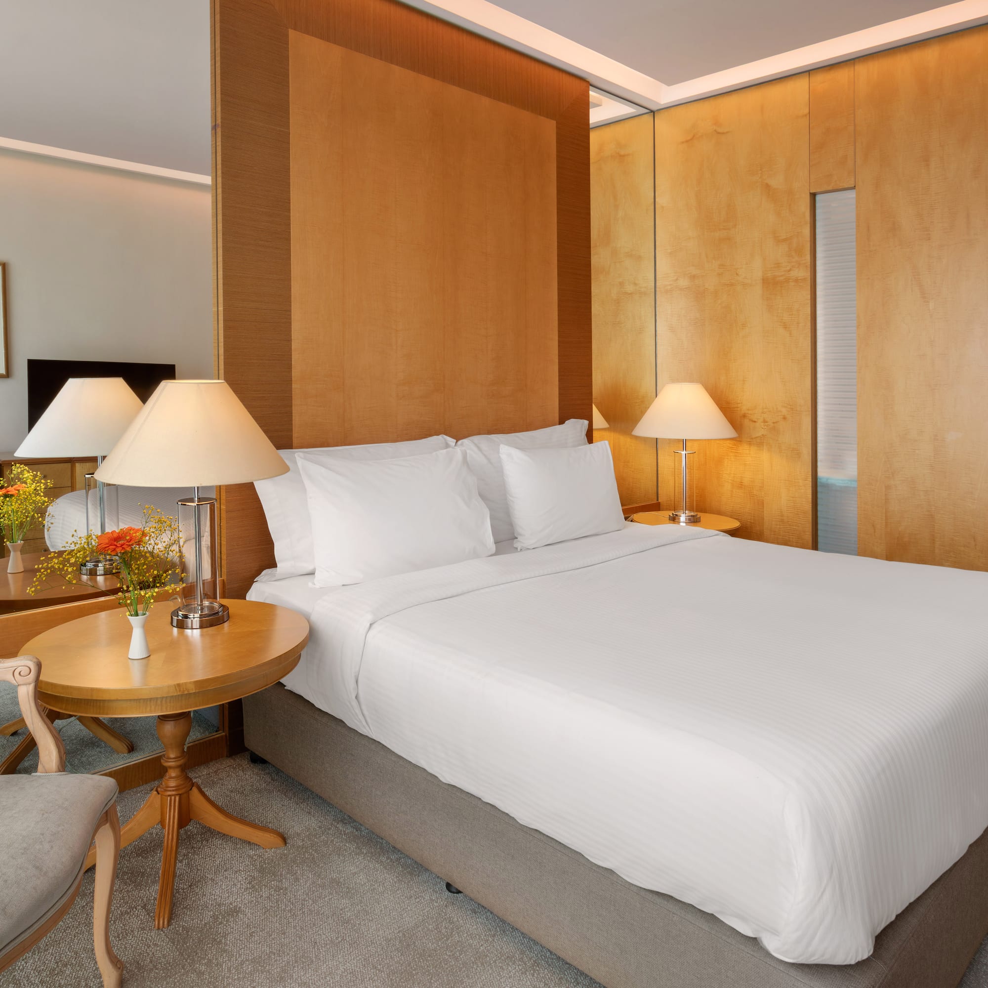 Elegant hotel bedroom with large bed, wooden panels, bedside lamps, and a vanity area.