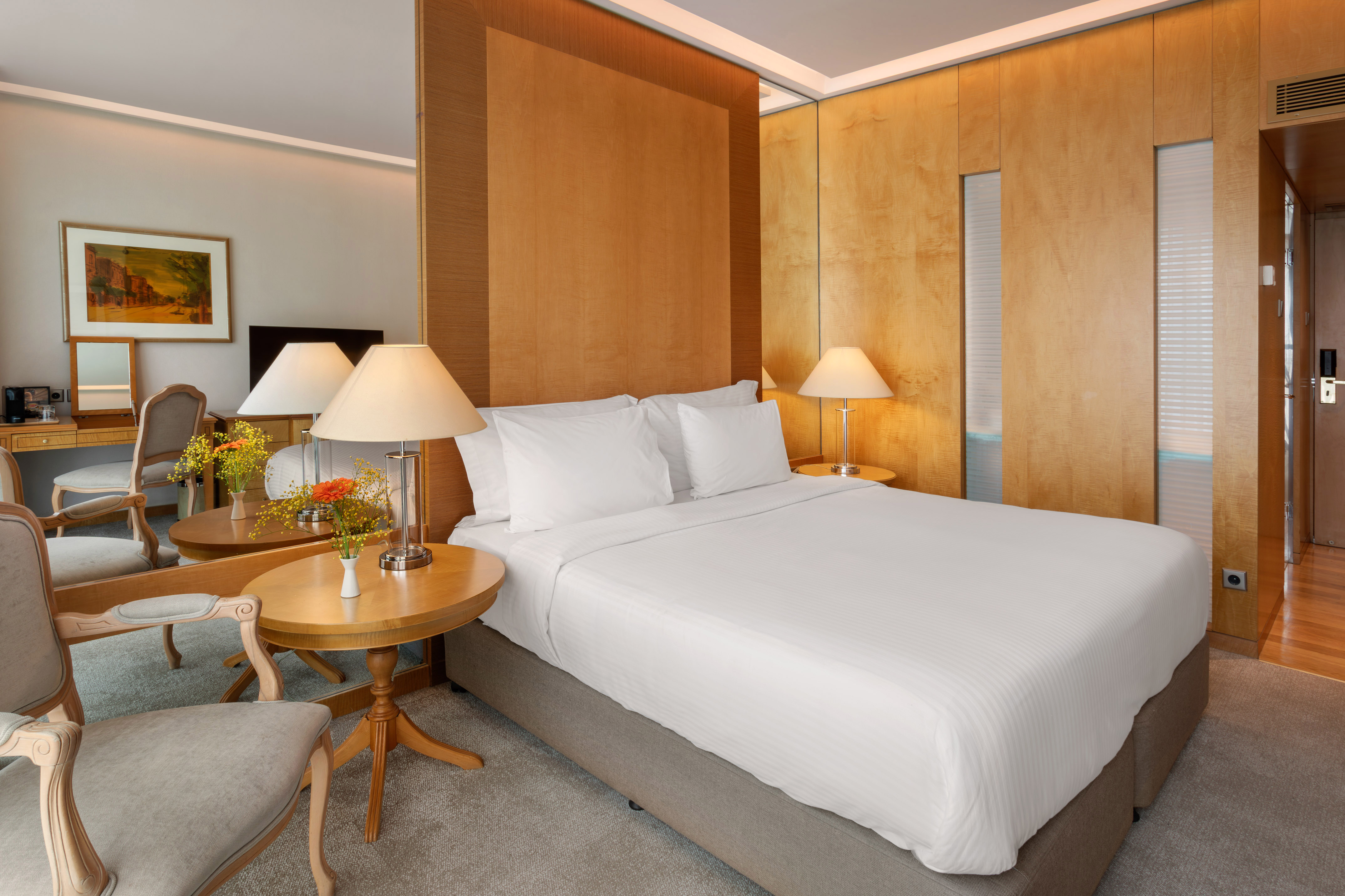 Elegant hotel bedroom with large bed, wooden panels, bedside lamps, and a vanity area.