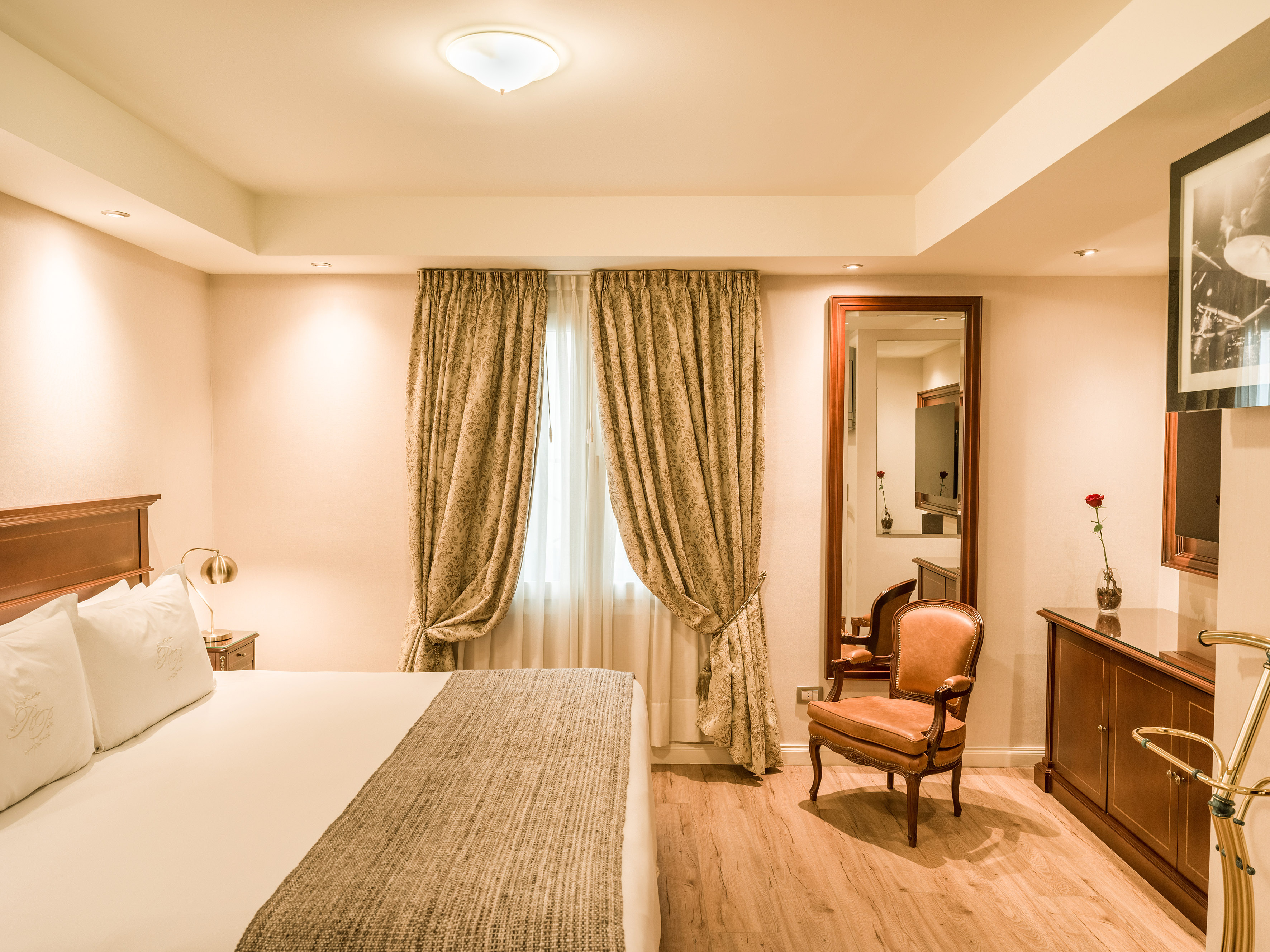 Elegant hotel room with king bed, ornate drapes, wood floors, and leather armchair.
