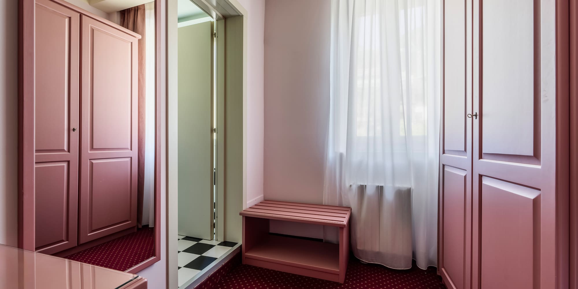 Rose pink bedroom with wardrobes, mirror, bench, and red patterned carpet.