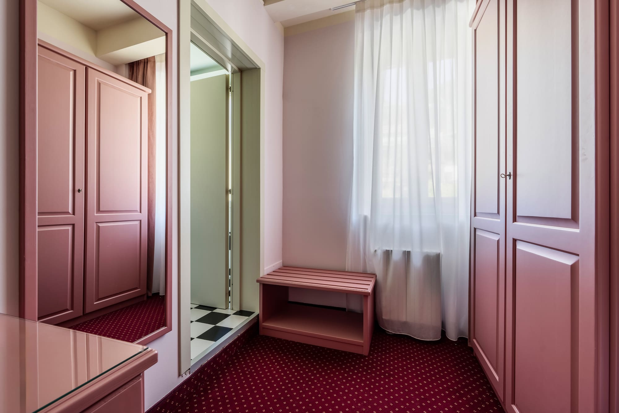 Rose pink bedroom with wardrobes, mirror, bench, and red patterned carpet.