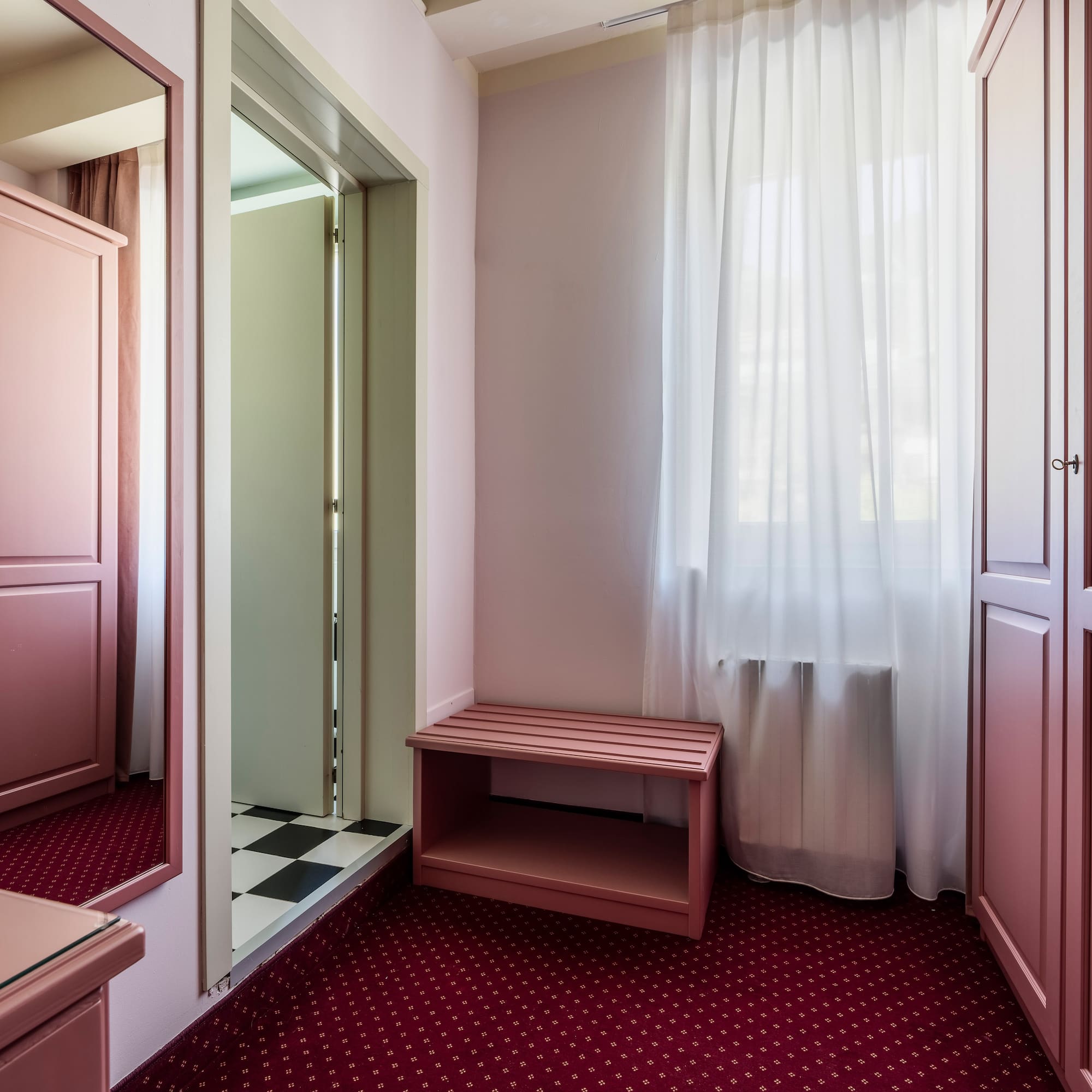 Rose pink bedroom with wardrobes, mirror, bench, and red patterned carpet.