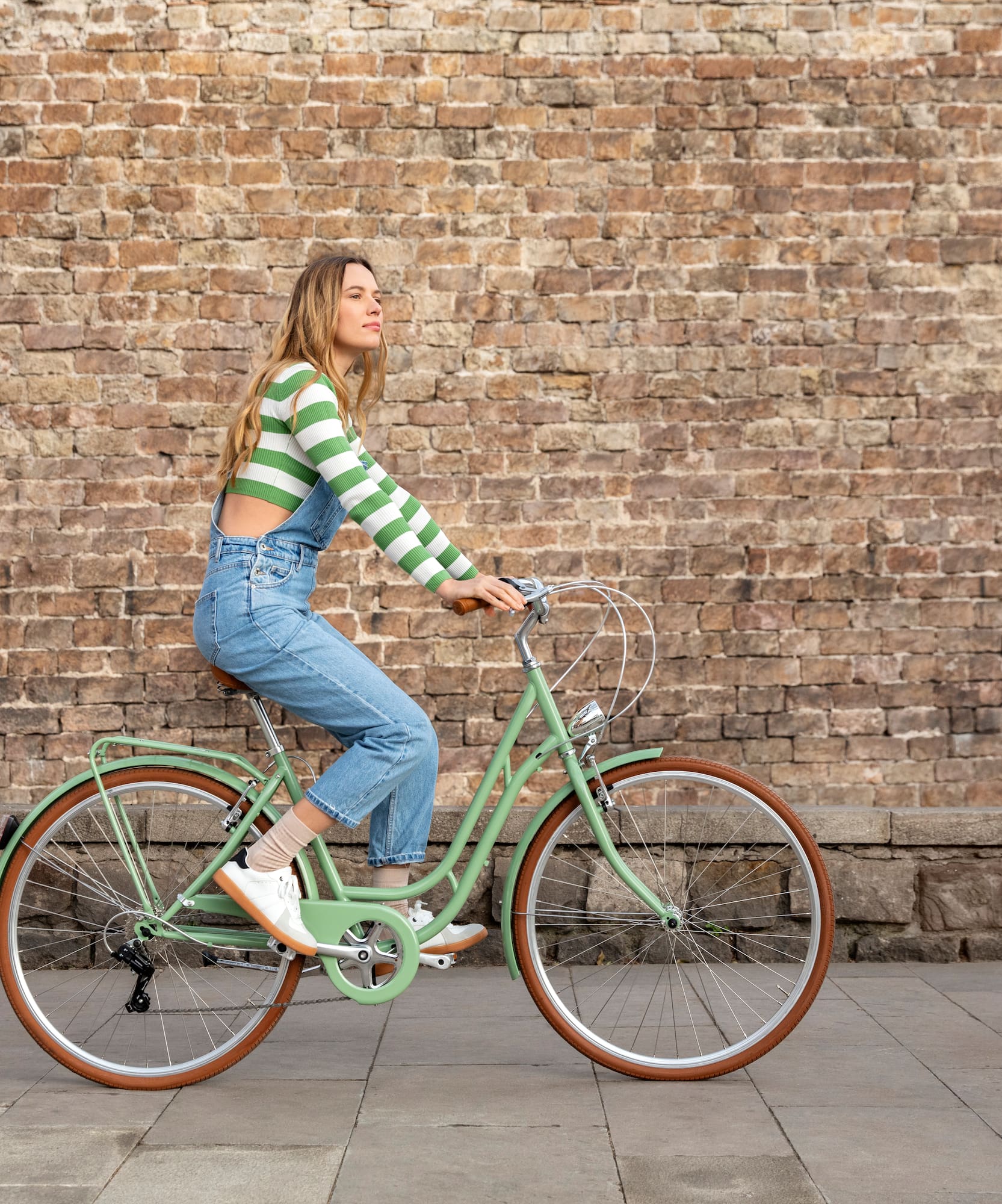 Mujer en bicicleta verde menta, mono, paseando junto a pared de ladrillo.