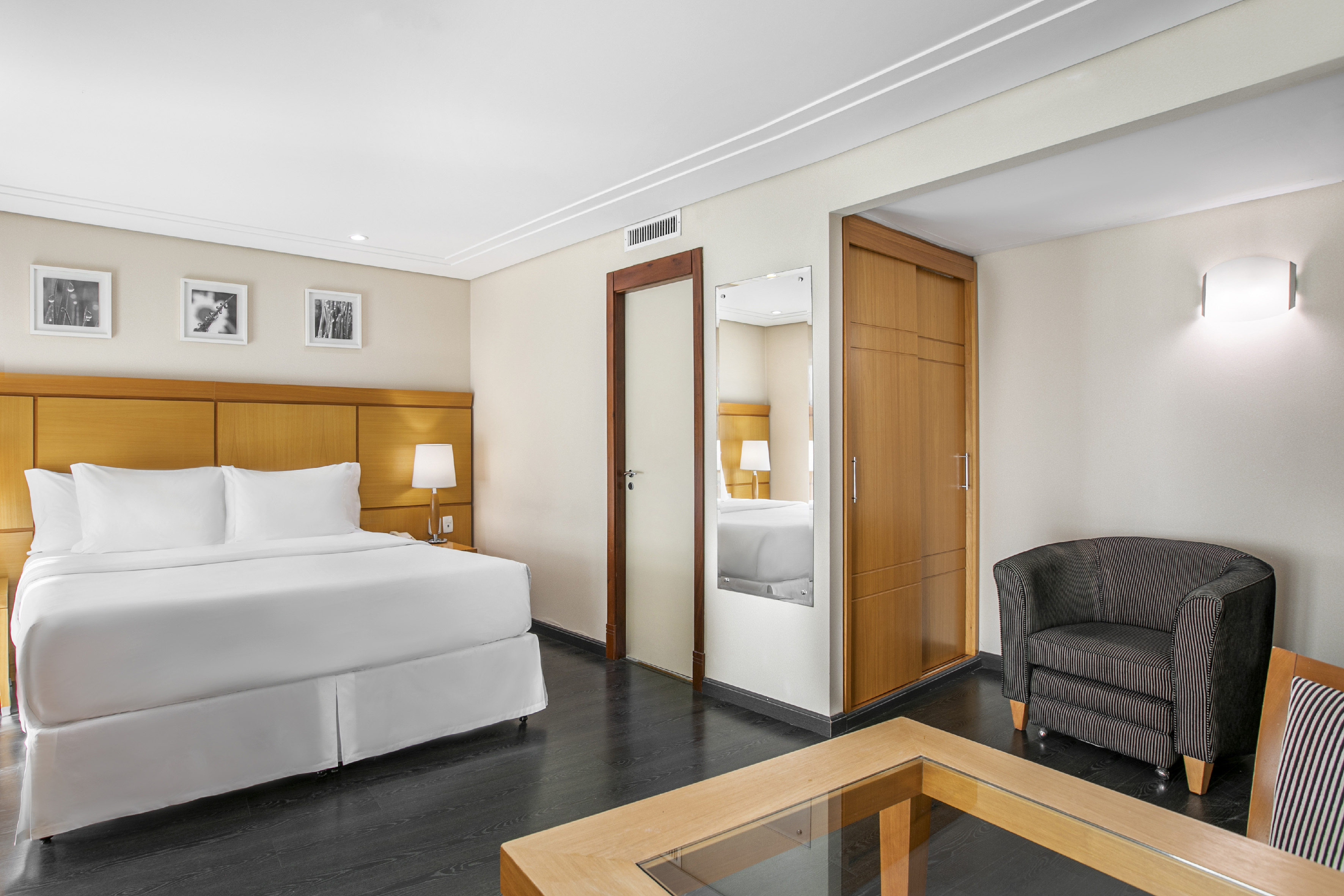 Modern hotel room with king bed, wooden furniture, striped armchair, and dark floor.