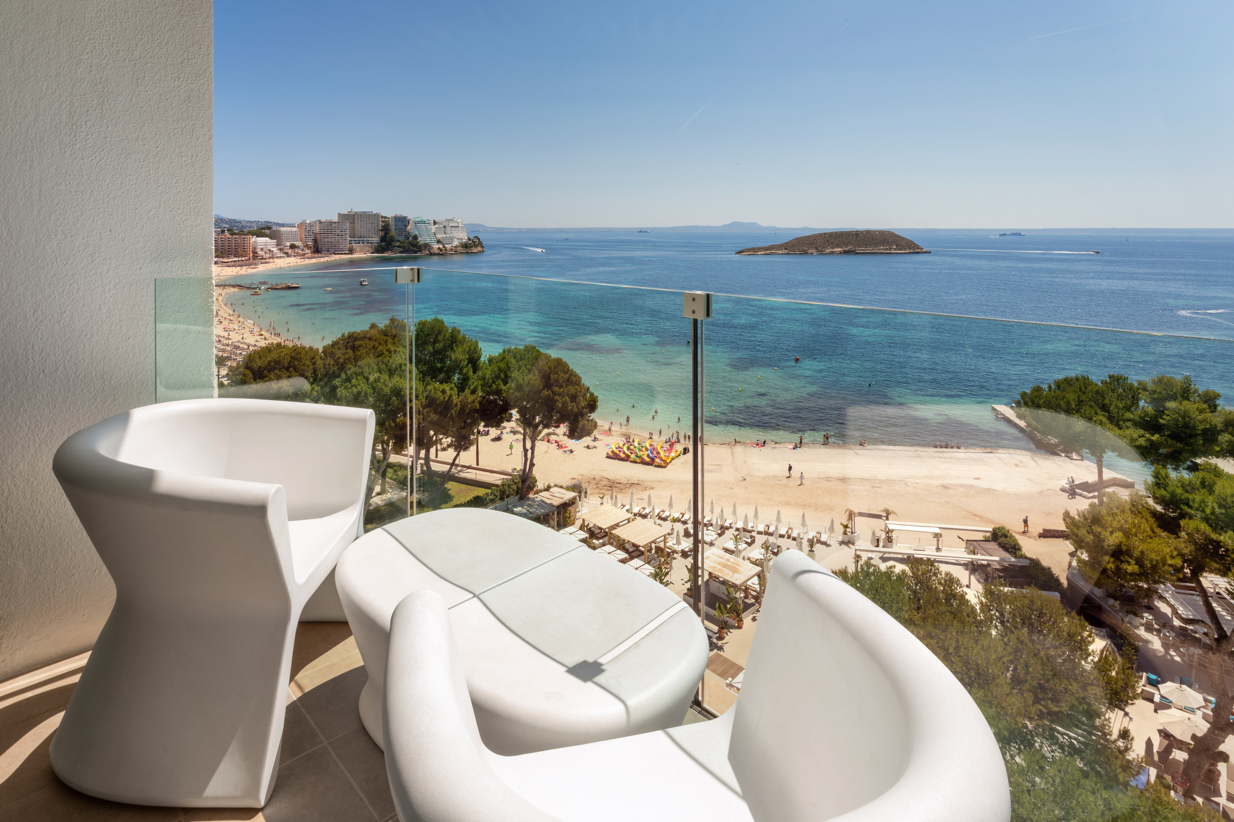 Modern balcony overlooking sunny beach, turquoise ocean, coastal resort buildings.