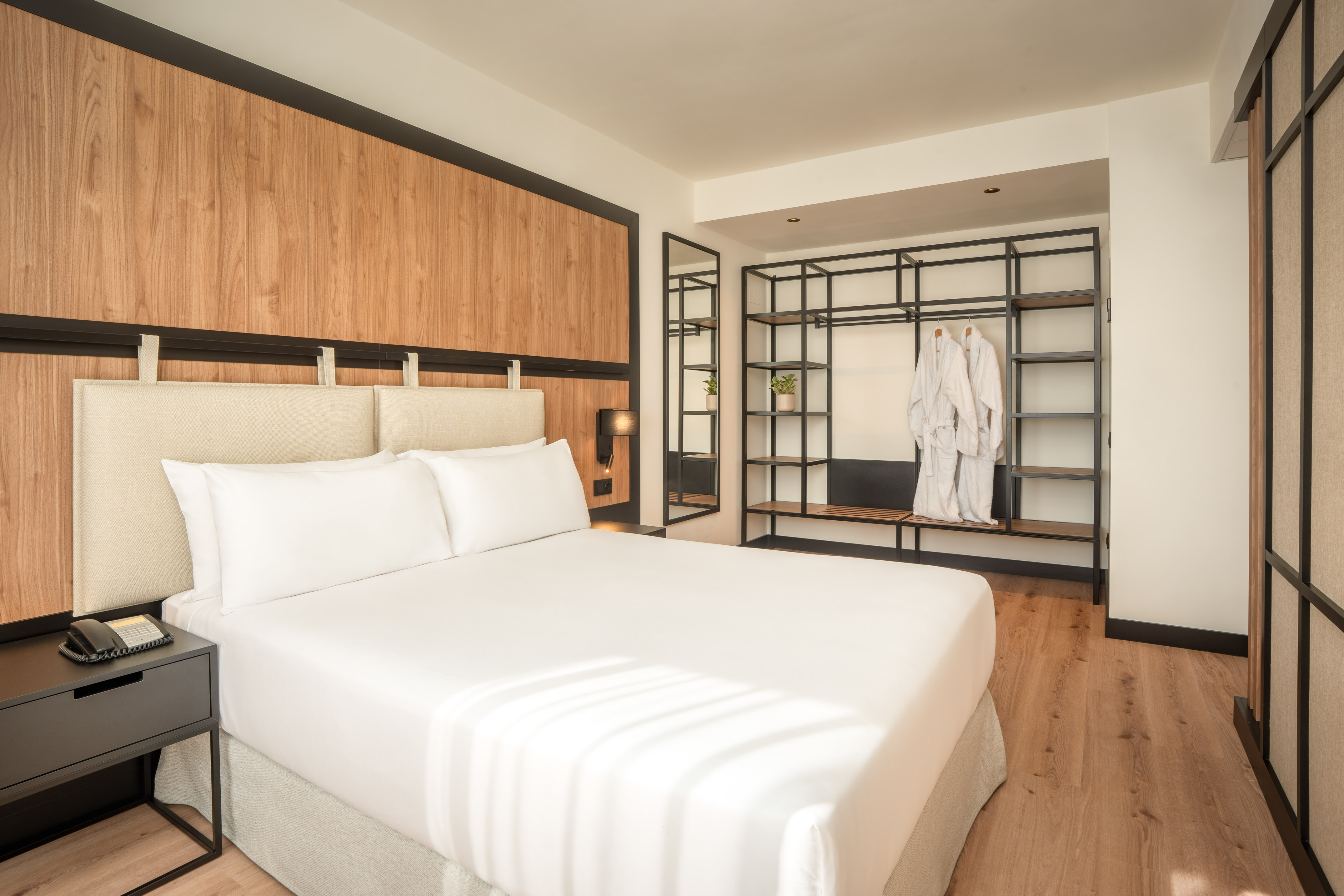 Hotel bedroom with modern white bed, wood wall, and open shelving with bathrobes.