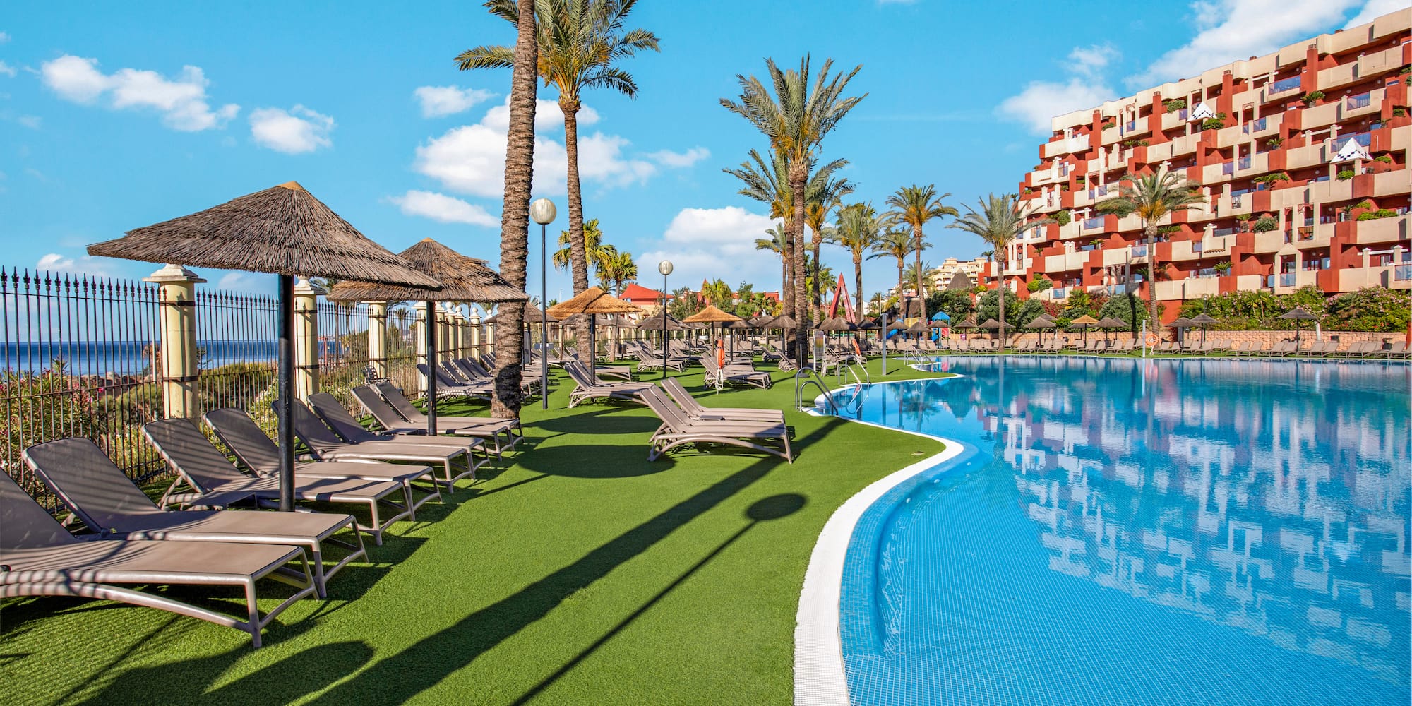 Sunny resort pool with lounge chairs, palm trees, hotel, and distant ocean view.