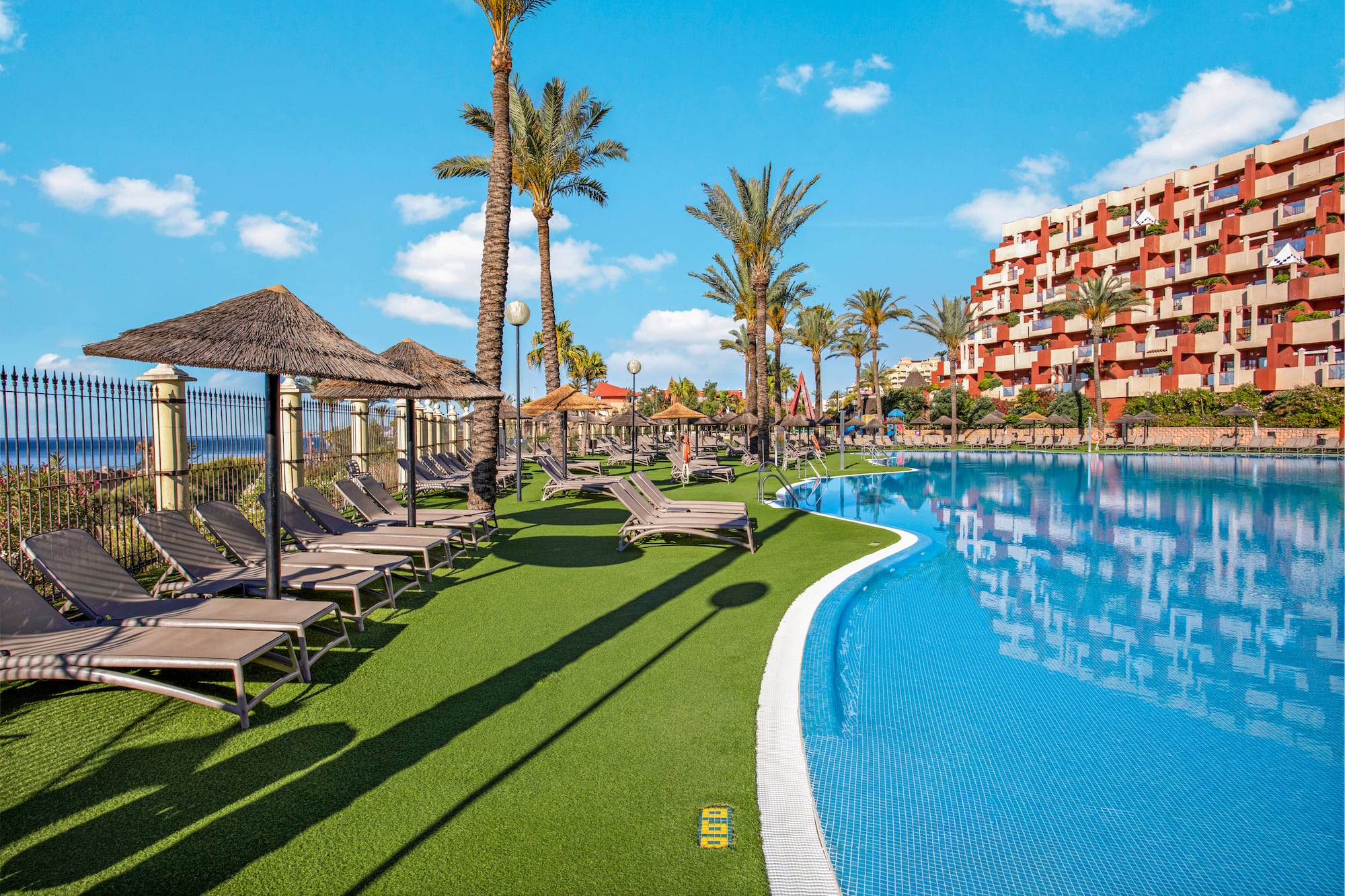 Sunny resort pool with lounge chairs, palm trees, hotel, and distant ocean view.