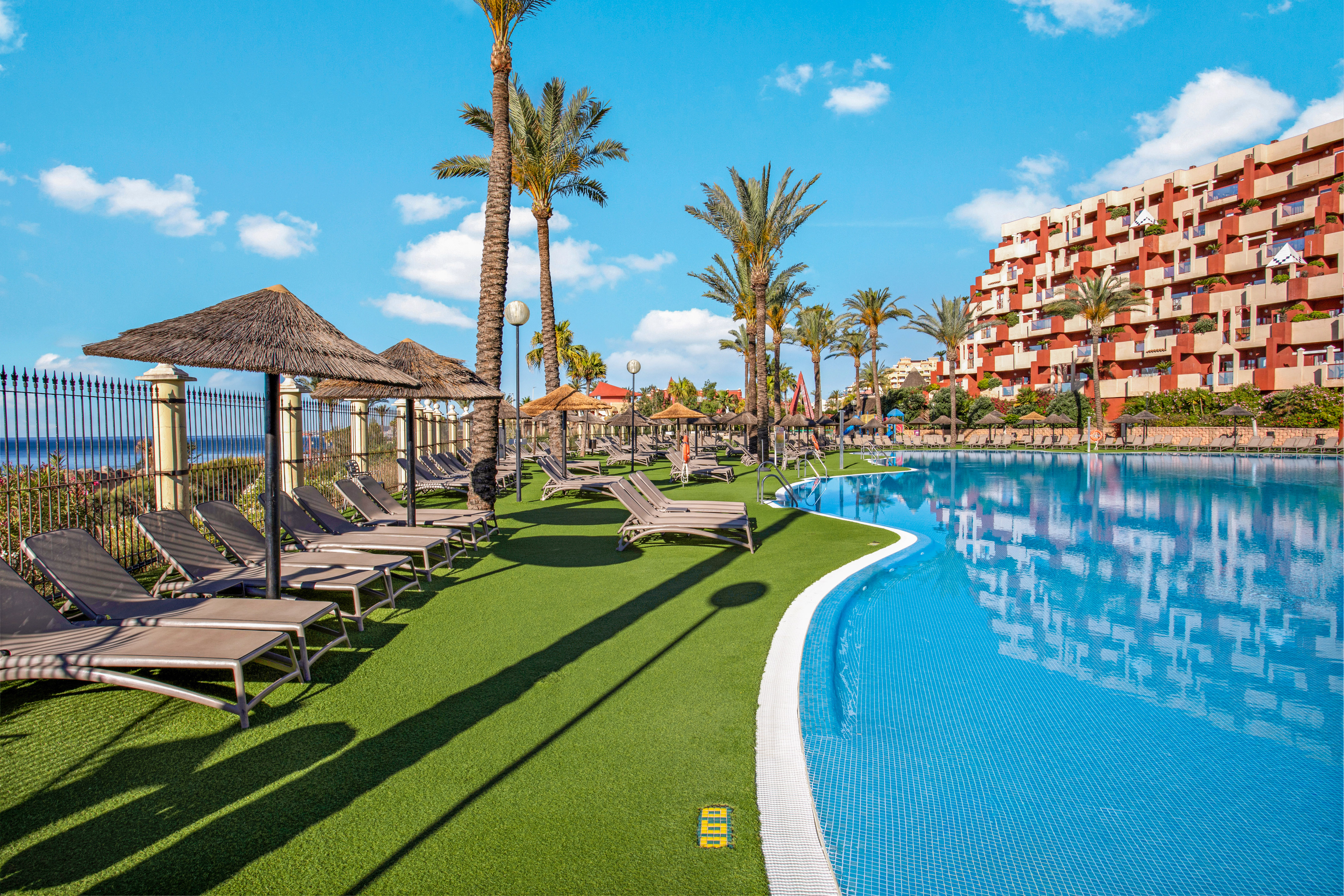 Sunny resort pool with lounge chairs, palm trees, hotel, and distant ocean view.