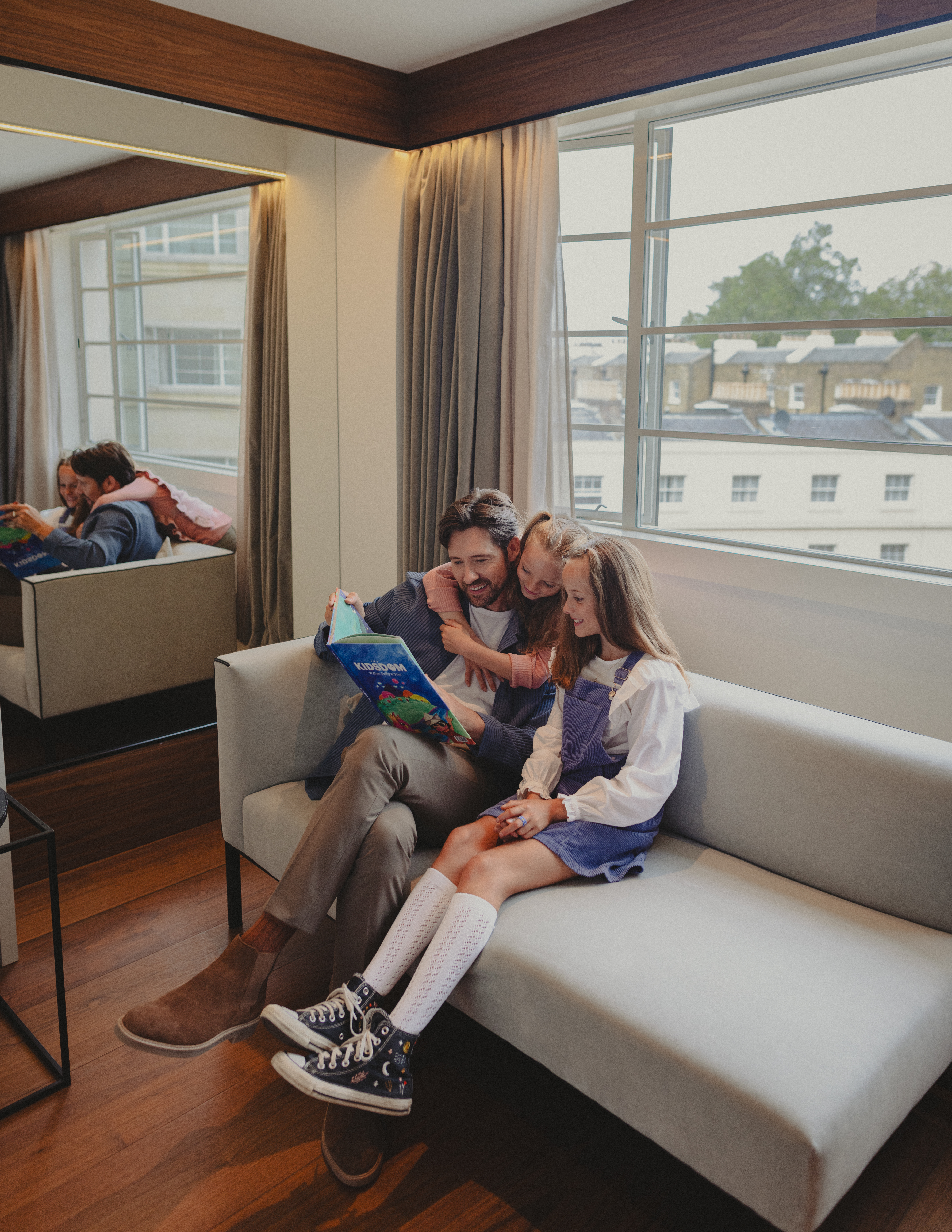 Father and two daughters reading a kids' book together on a cozy hotel sofa.