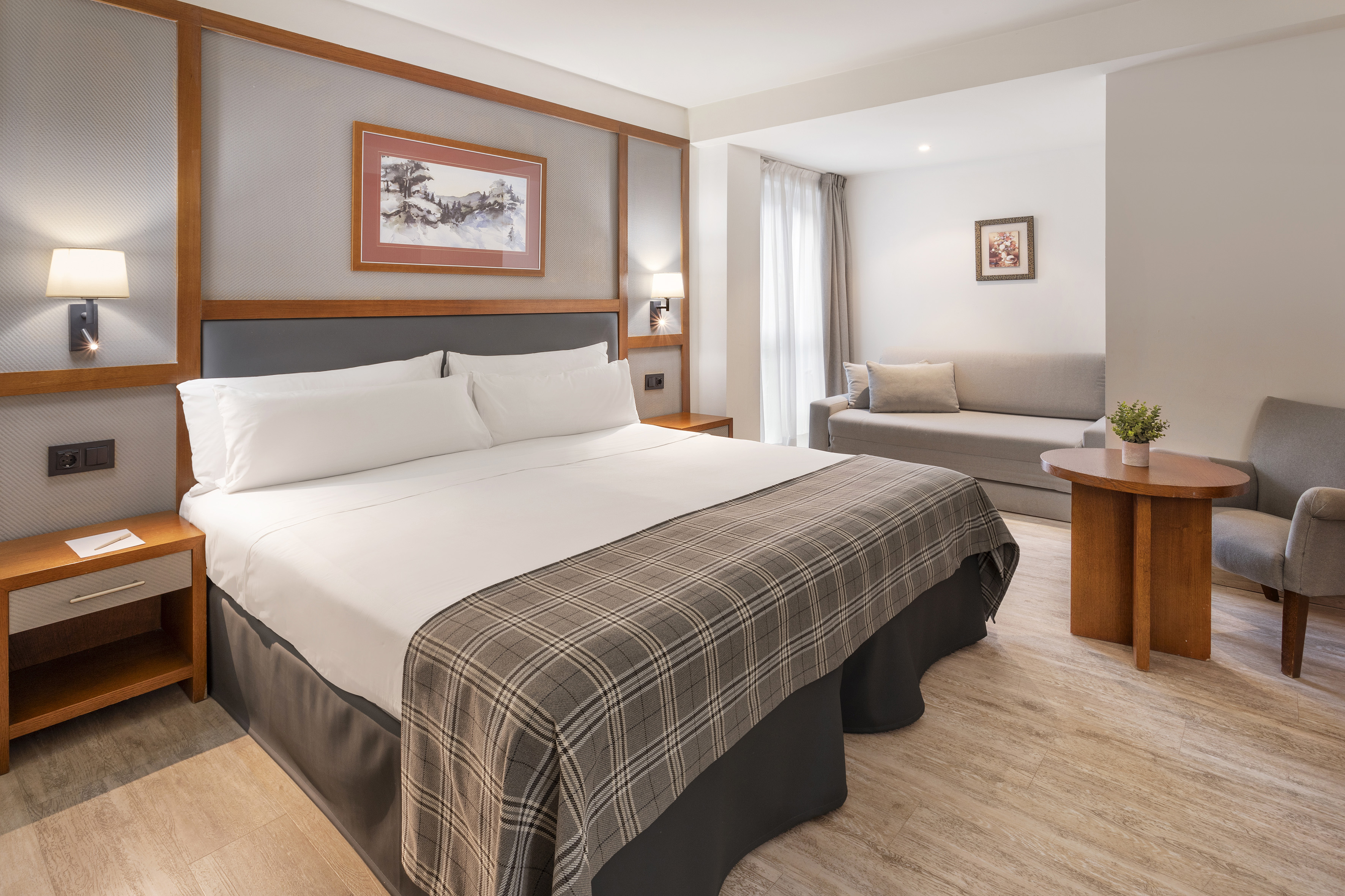 Modern hotel room with king bed, sofa, plaid blanket, and wooden accents.