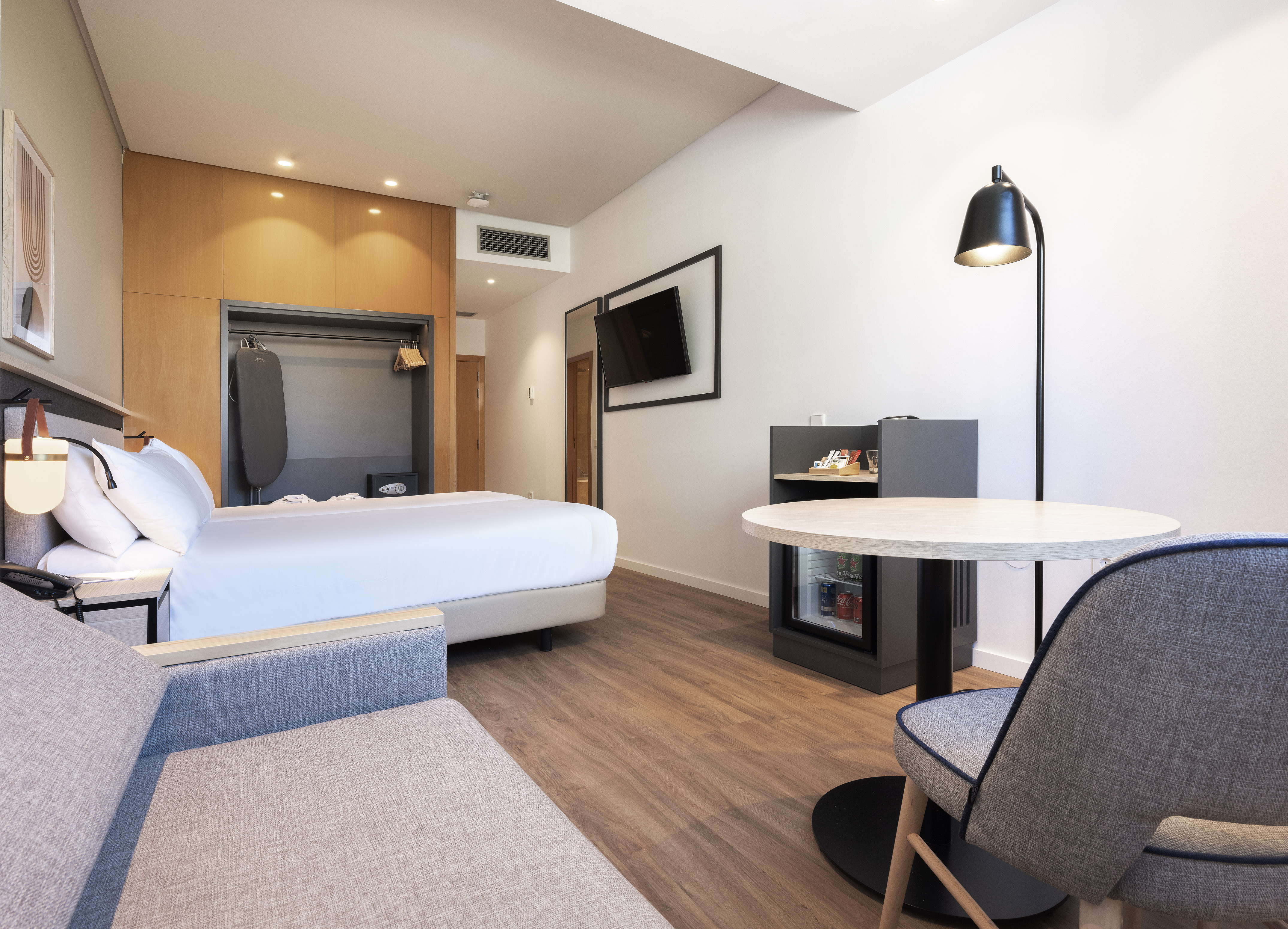 Modern hotel room with bed, gray sofa, wooden closet, TV, and round table.