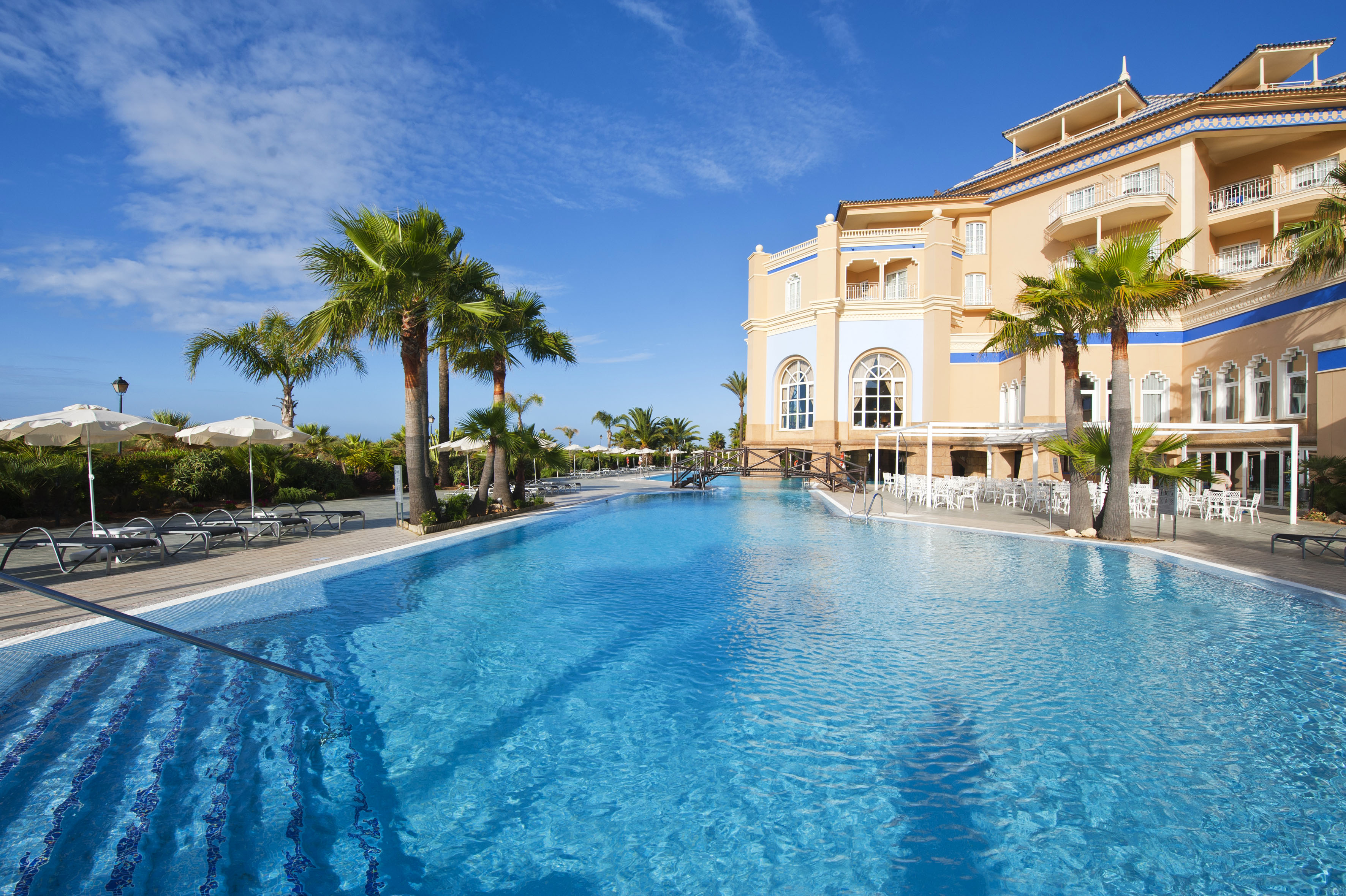 Large resort pool, palm trees, and Mediterranean hotel on a sunny day.